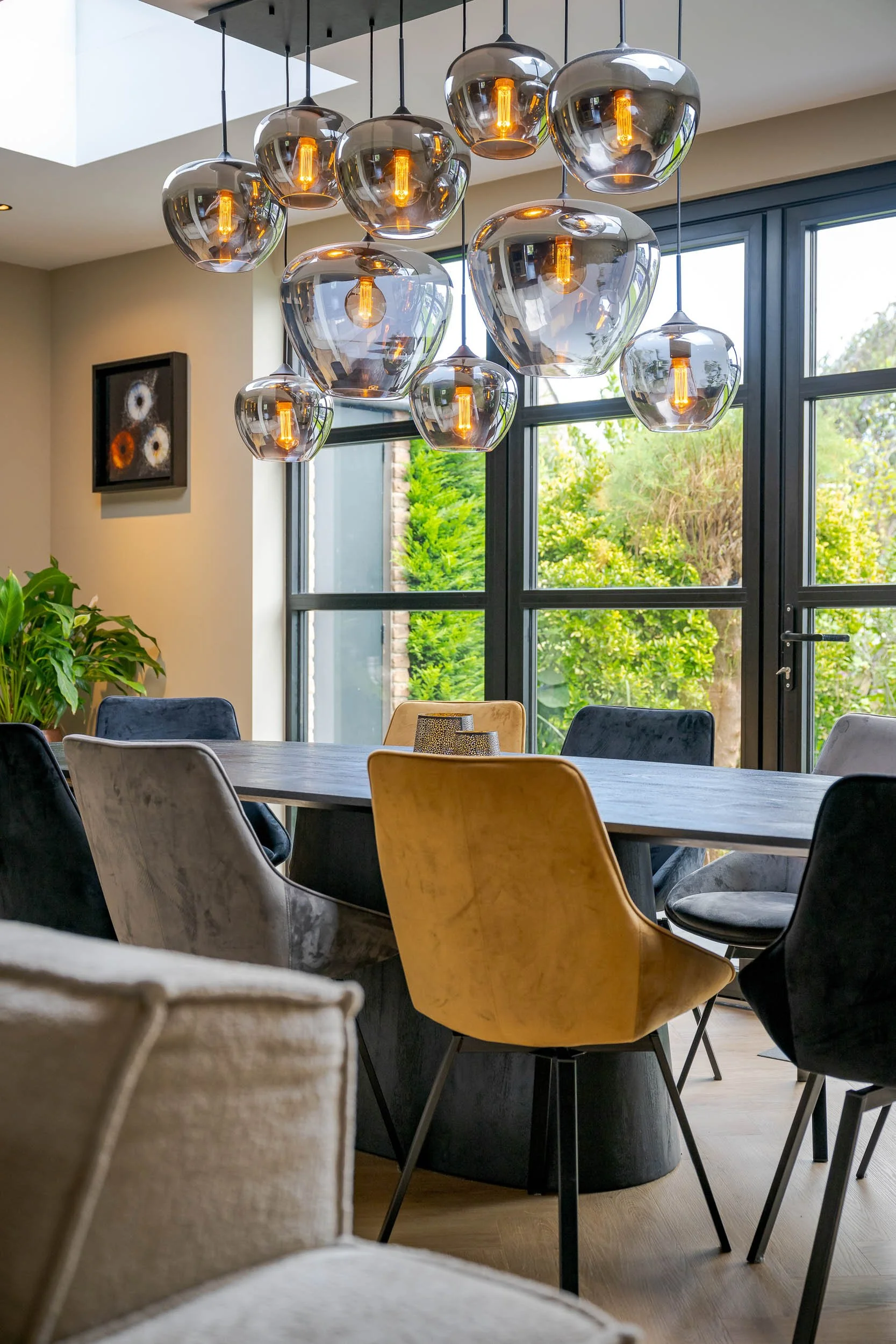 Een moderne eetkamer met een grote tafel, diverse stoelen, grote ramen die uitzicht bieden op groene tuin, en een verlichte, glazen hanglamp met meerdere kappen.