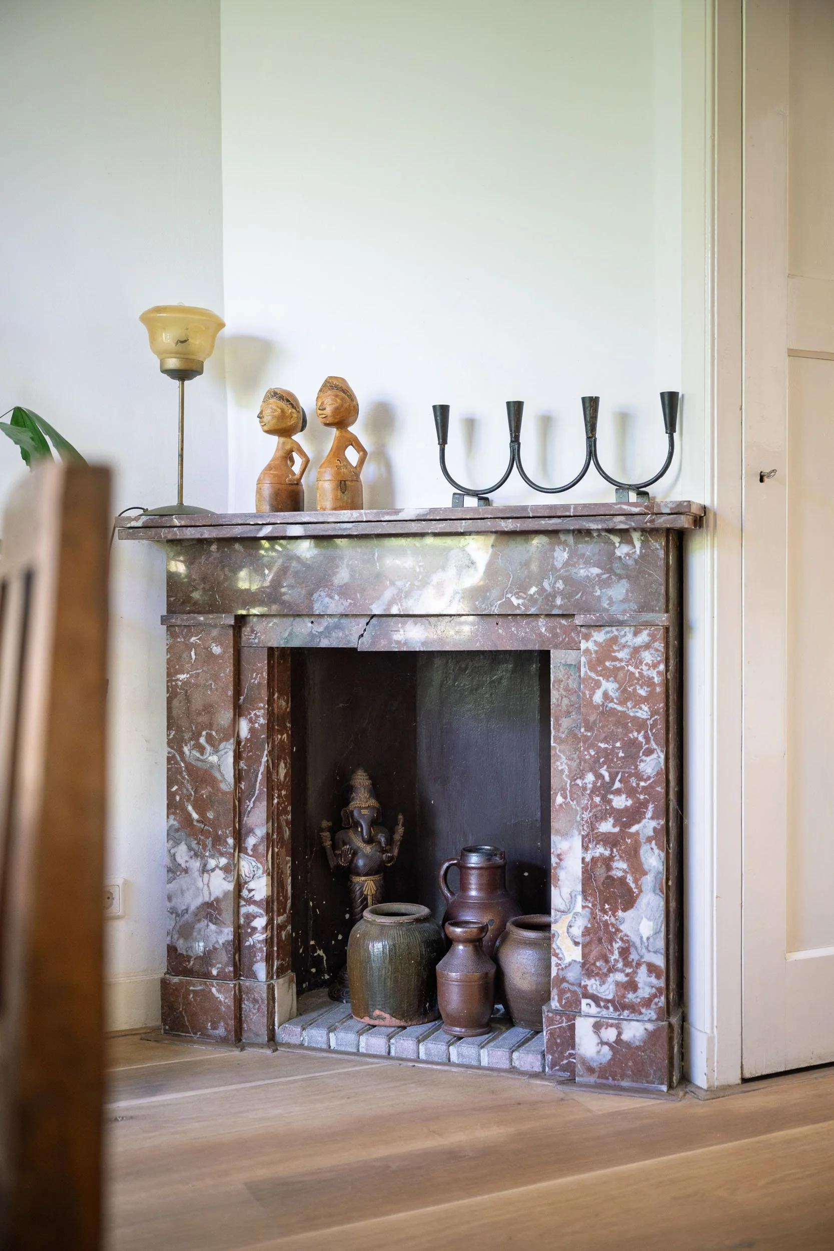 Een open haard met een rode marmeren mantel, decoratief aardewerk en beelden binnen in een huis, met enkele vazen en een beeld in de opening van de haard.