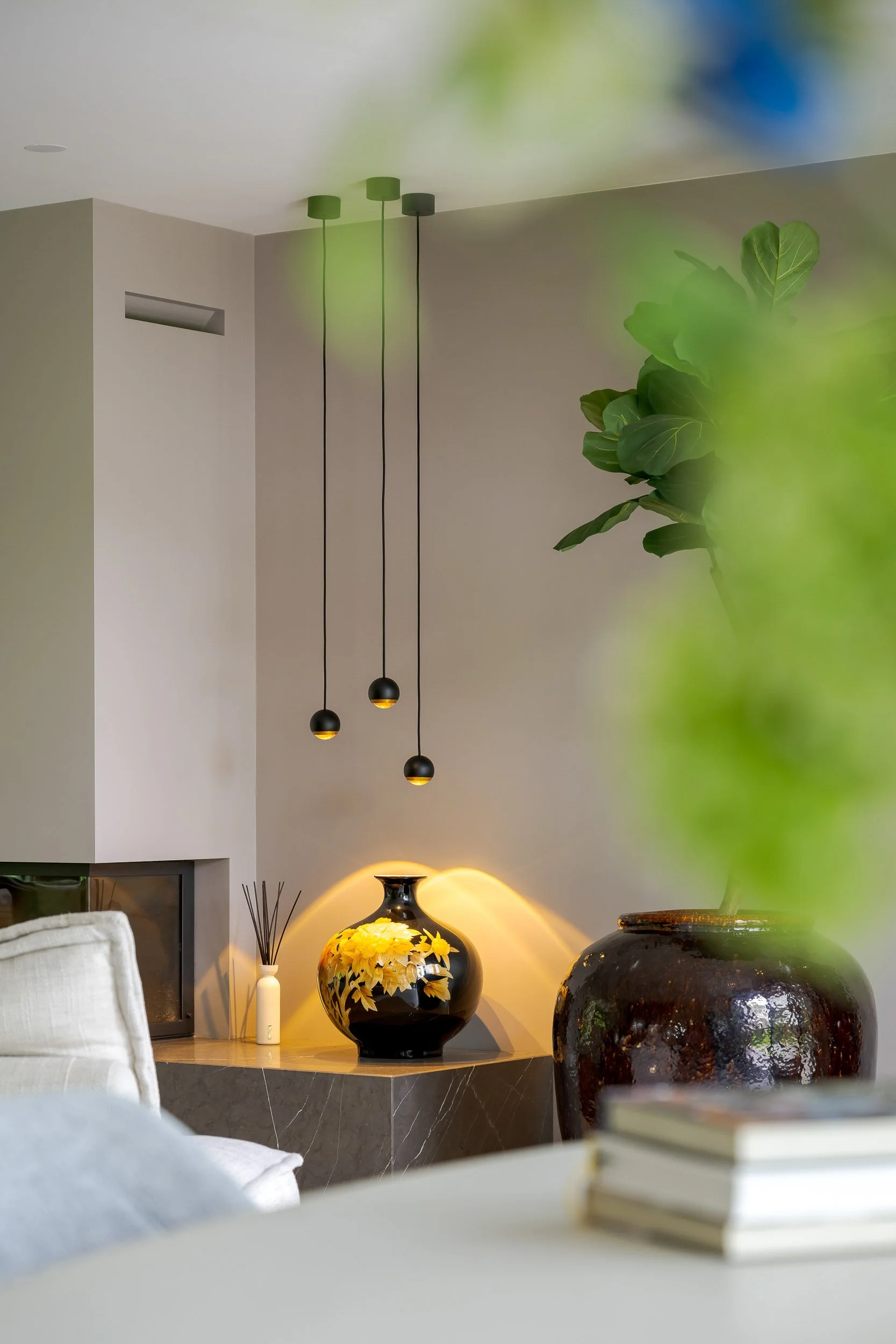 Een interieur met hangende lichtlampen, een grote groene plant, een zwart ornament met gele bloemendecoratie, en decoratieve objecten op een tafel.