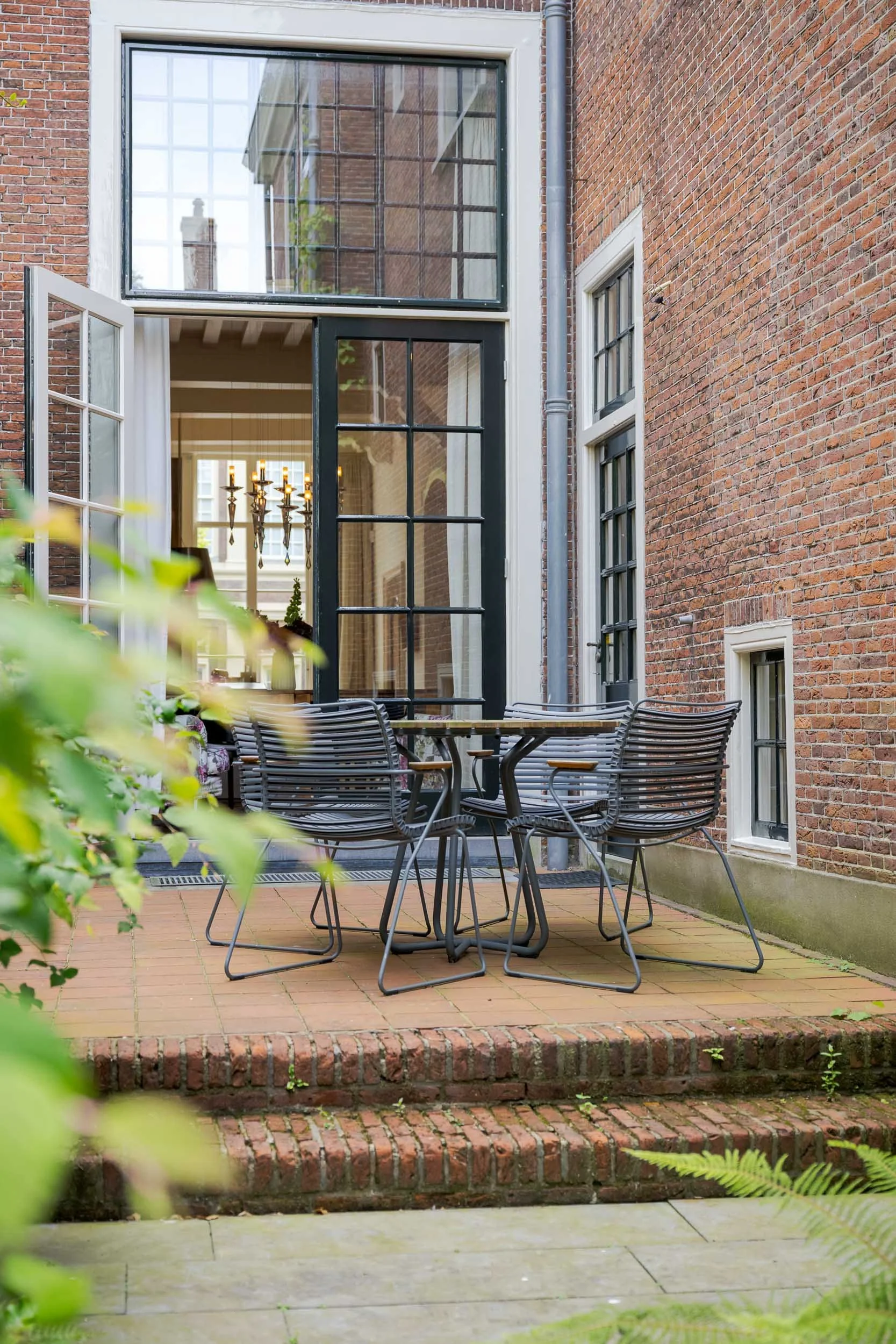 Buiten patio met metalen tafel en stoelen, omgeven door rode bakstenen muren en een grotere glazen deur en ramen, met planten op de rand en felle verlichting binnen.