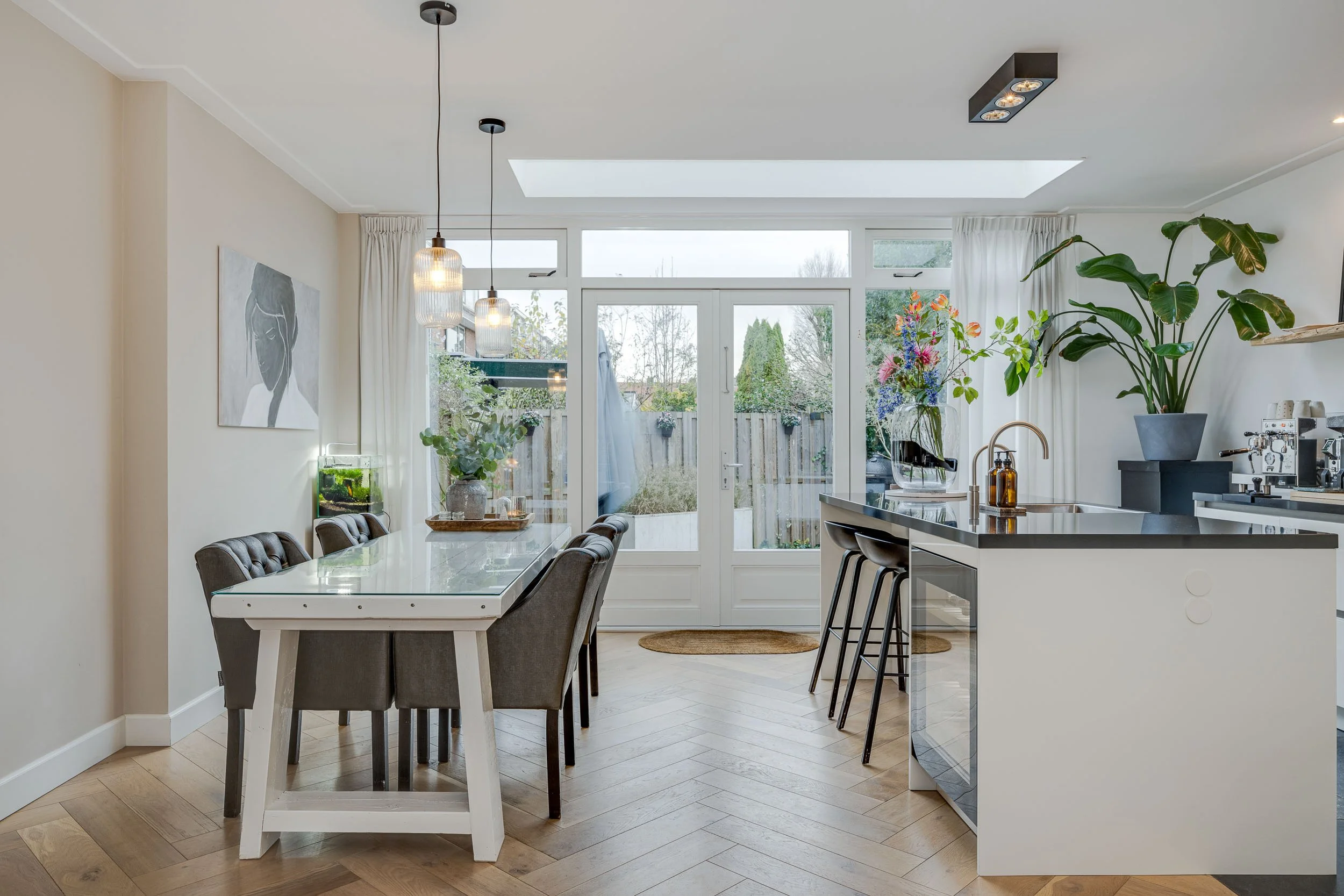 De afbeelding toont een moderne, lichte keuken en eetkamer met grote ramen en een glasdeur die uitzicht bieden op de tuin. Er hangen twee hanglampen boven de eettafel. Op de tafel staan vazen met bloemen en een klein aquarium. Aan de rechterkant staa