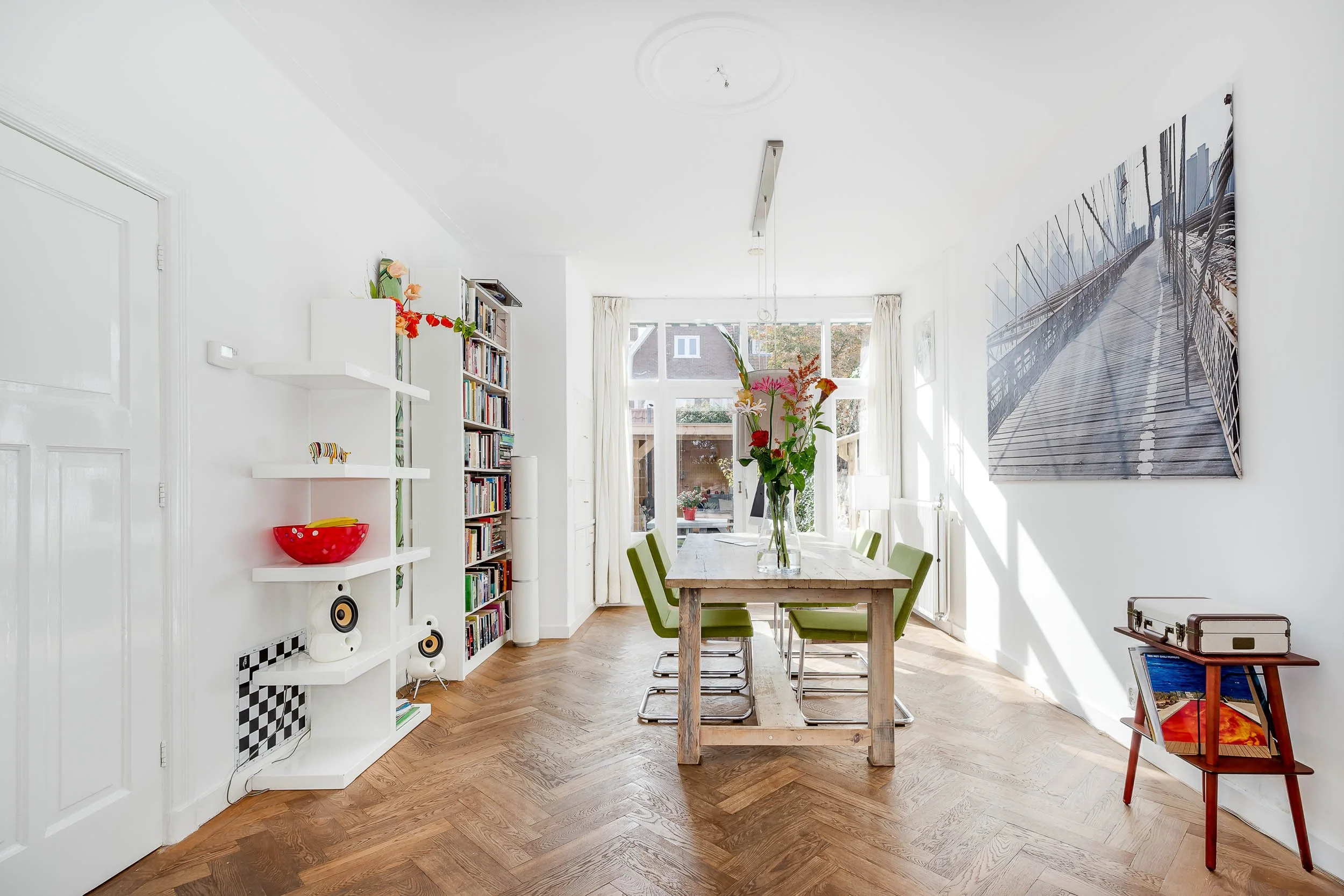 Een lichte eetkamer met een houten tafel en zes groene stoelen. Aan de muur hangt een groot zwart-wit foto van een brug. Er staan bloemen op de tafel. In de achtergrond grote ramen met uitzicht op een tuin.