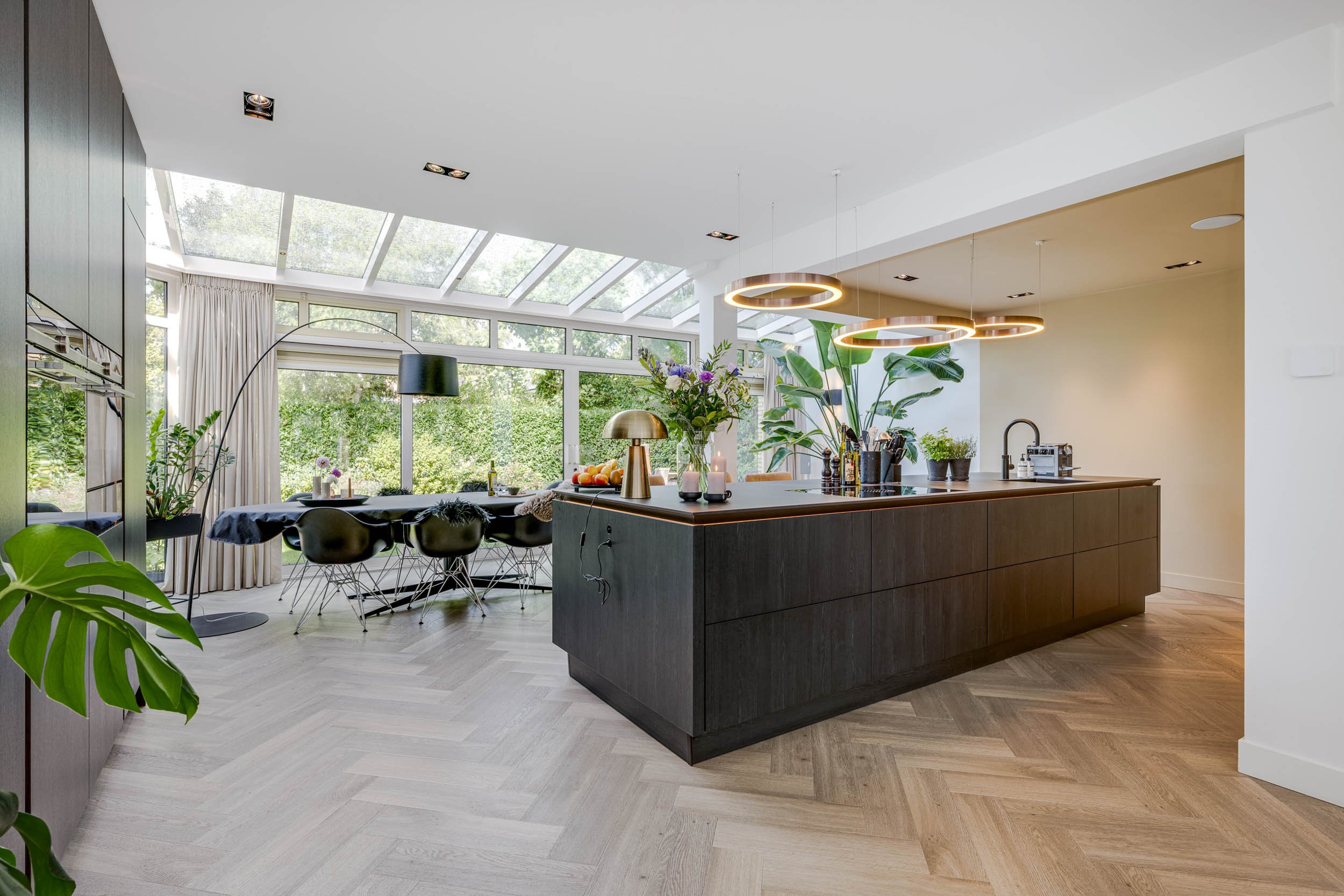 Een moderne keuken met een groot breakfast bar, groene planten, ronde hanglampen, grote ramen en veel natuurlijk licht, met buitenzicht op een tuin.