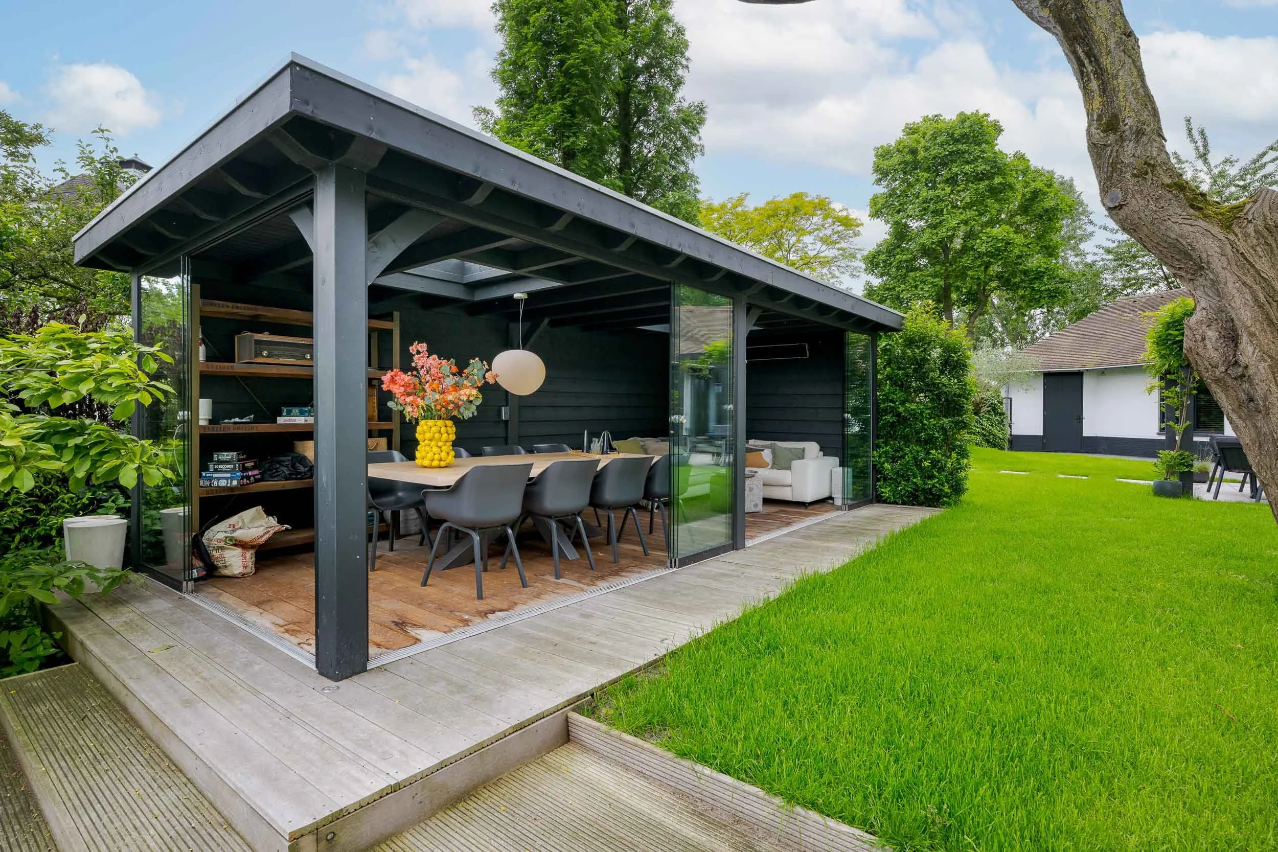 Een moderne buitenterrasoverkapping met glazen schuifdeuren, een eettafel met stoelen, een grote vaas met bloemen, een hanglamp en een zitbank met kussens in een groene tuin.