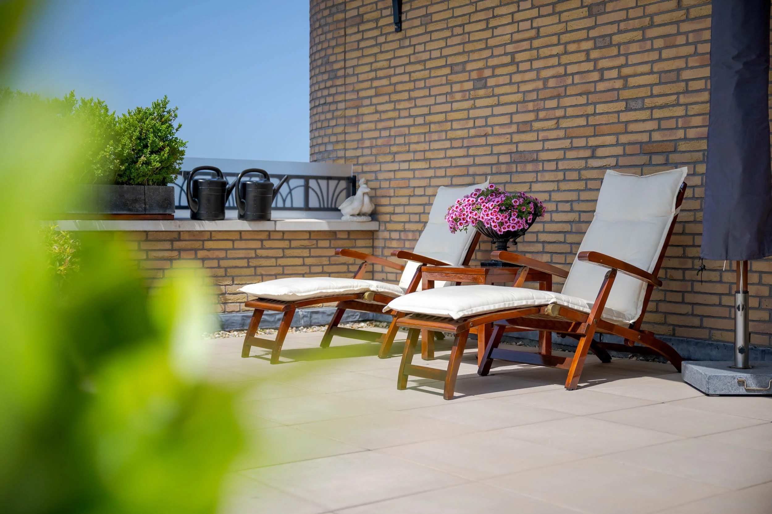 Twee ligstoel met witte kussens, pot met roze bloemen, een paar tuinaccessoires en planten op een balkon met een bakstenen muur.