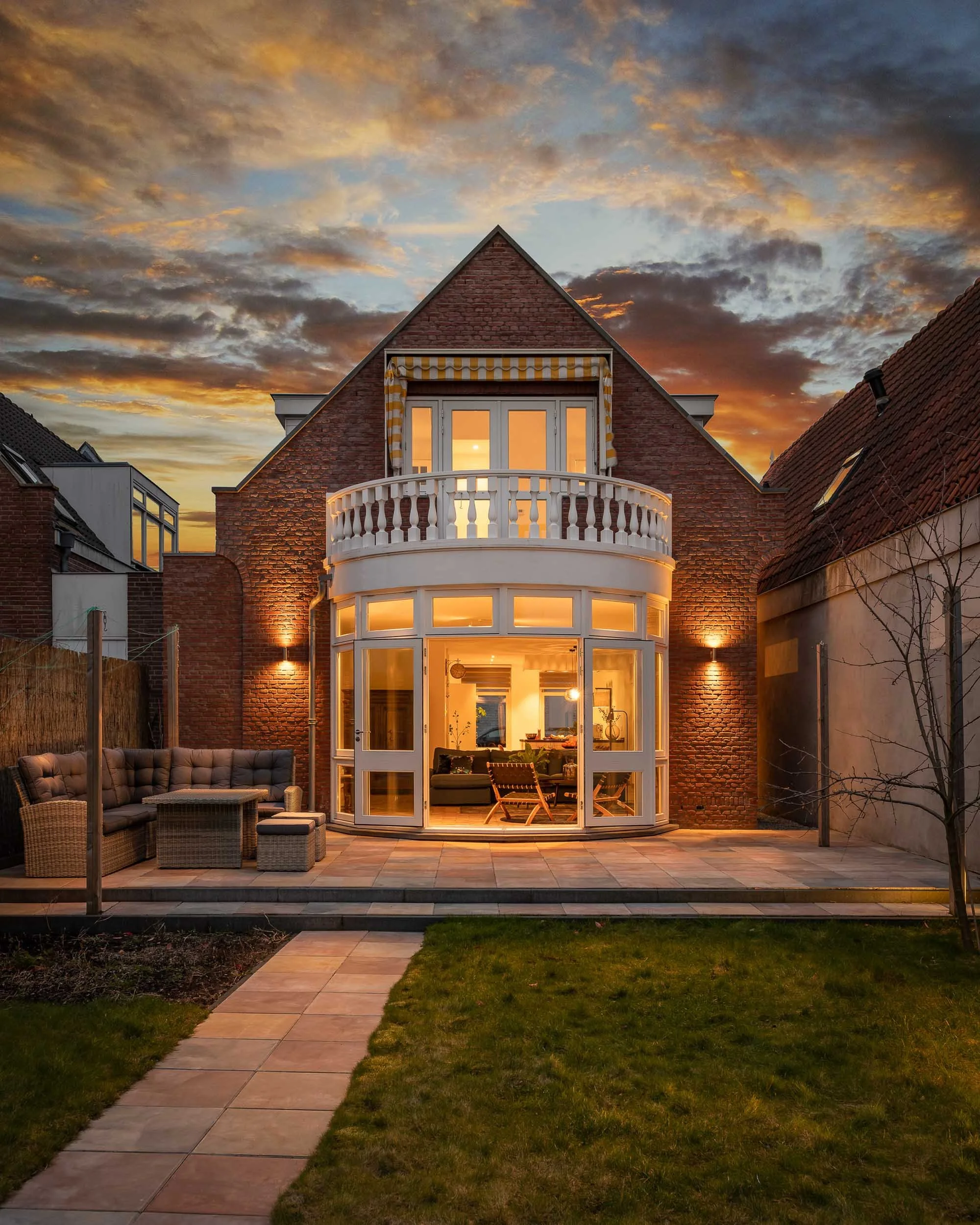 Een afgebeelde tuin met een patio, houten tuinmeubelen, een erf met groen gras en tegels, een achtertuin van een huis met een serre en een balkon en een schemerende hemel met oranje en blauwe wolken.