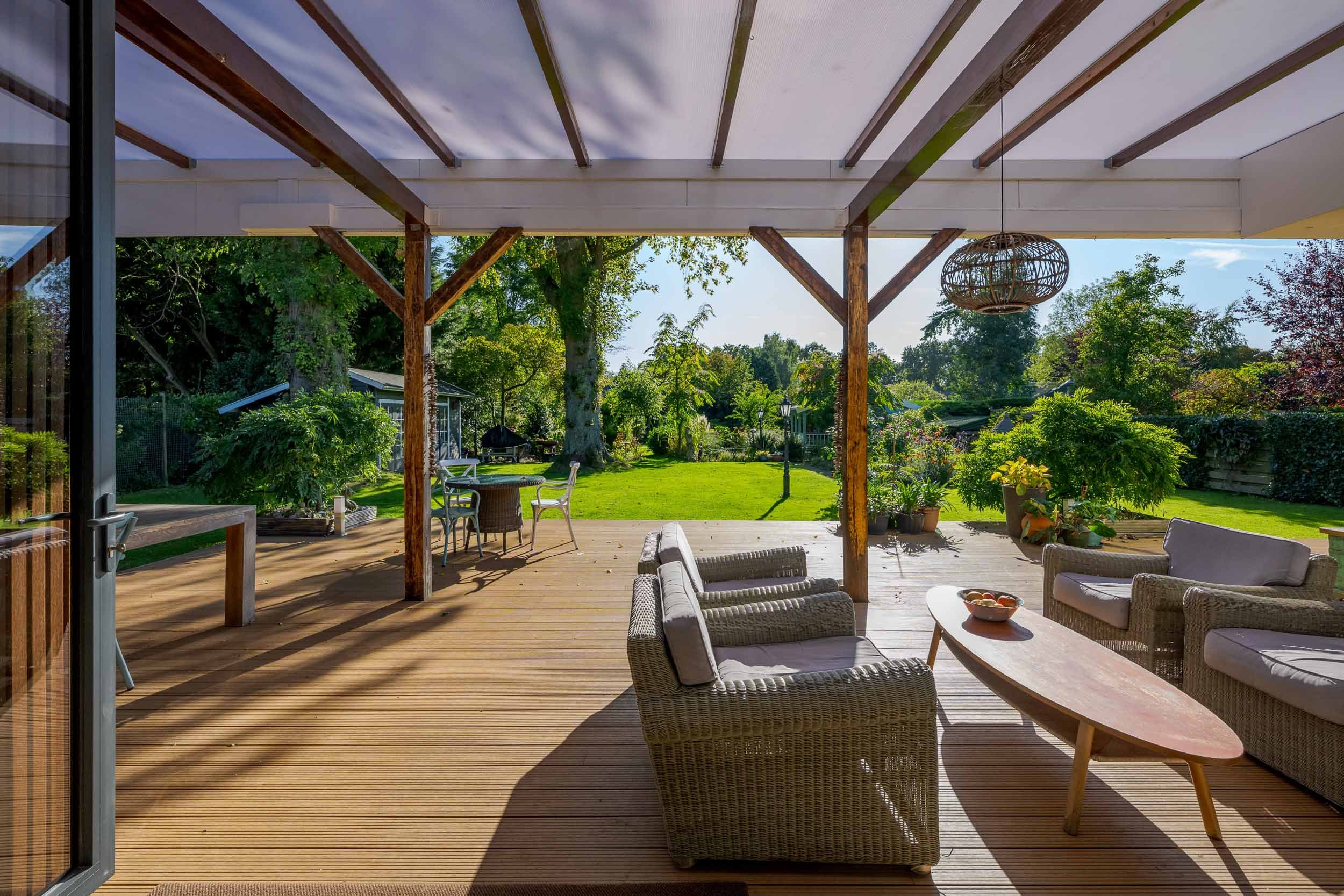 Uitzicht vanaf een gezellig houten terras met loungebed, eetkamerset en uitzicht op een groene tuin met bomen en gras onder een overkapping.