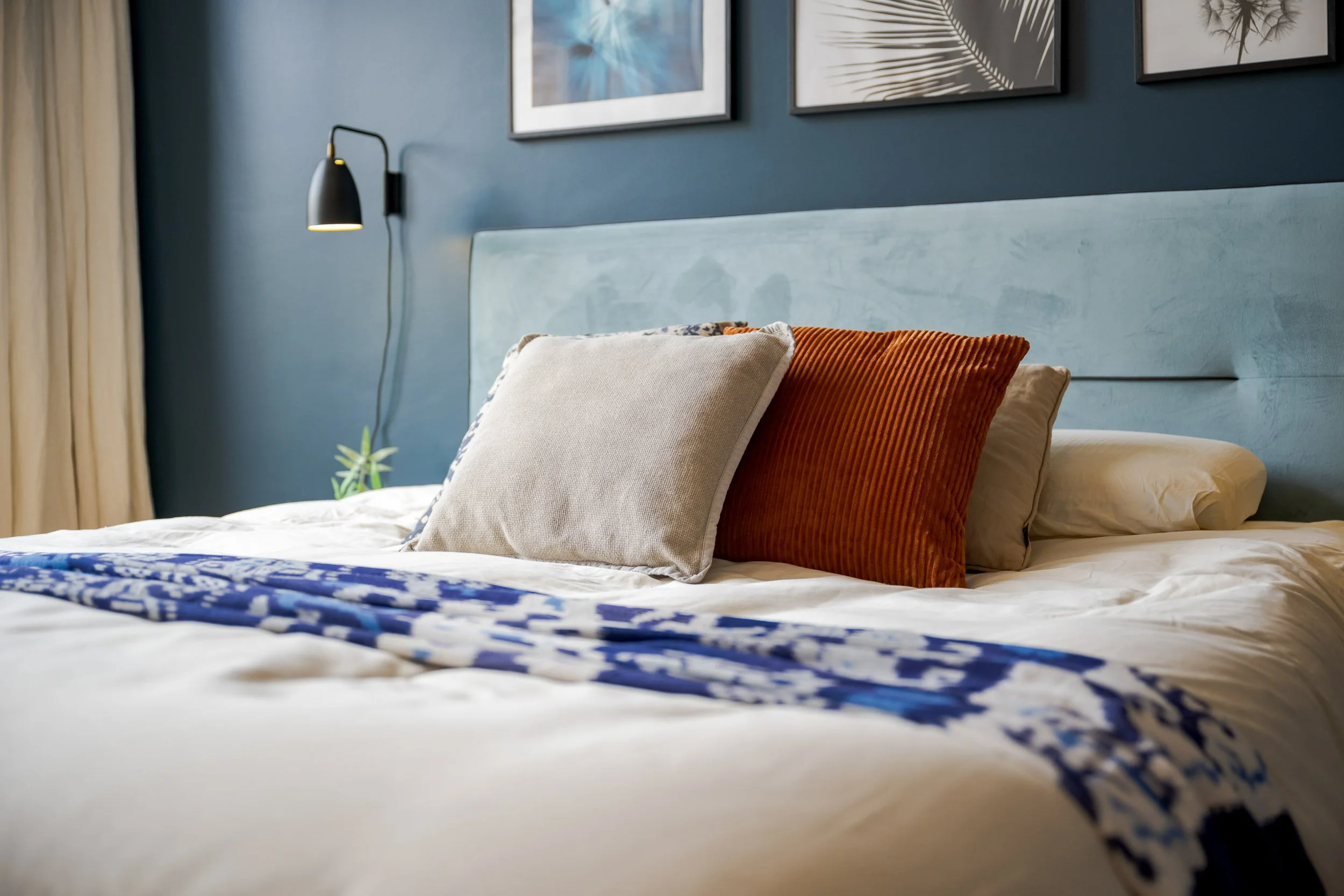 Een slaapkamer met blauwe muur, betegeld hoofdeinde, drie framed prints met botanische ontwerpen, en een bed met kussens in beige en oranje tinten, beddengoed met blauwe patronen.