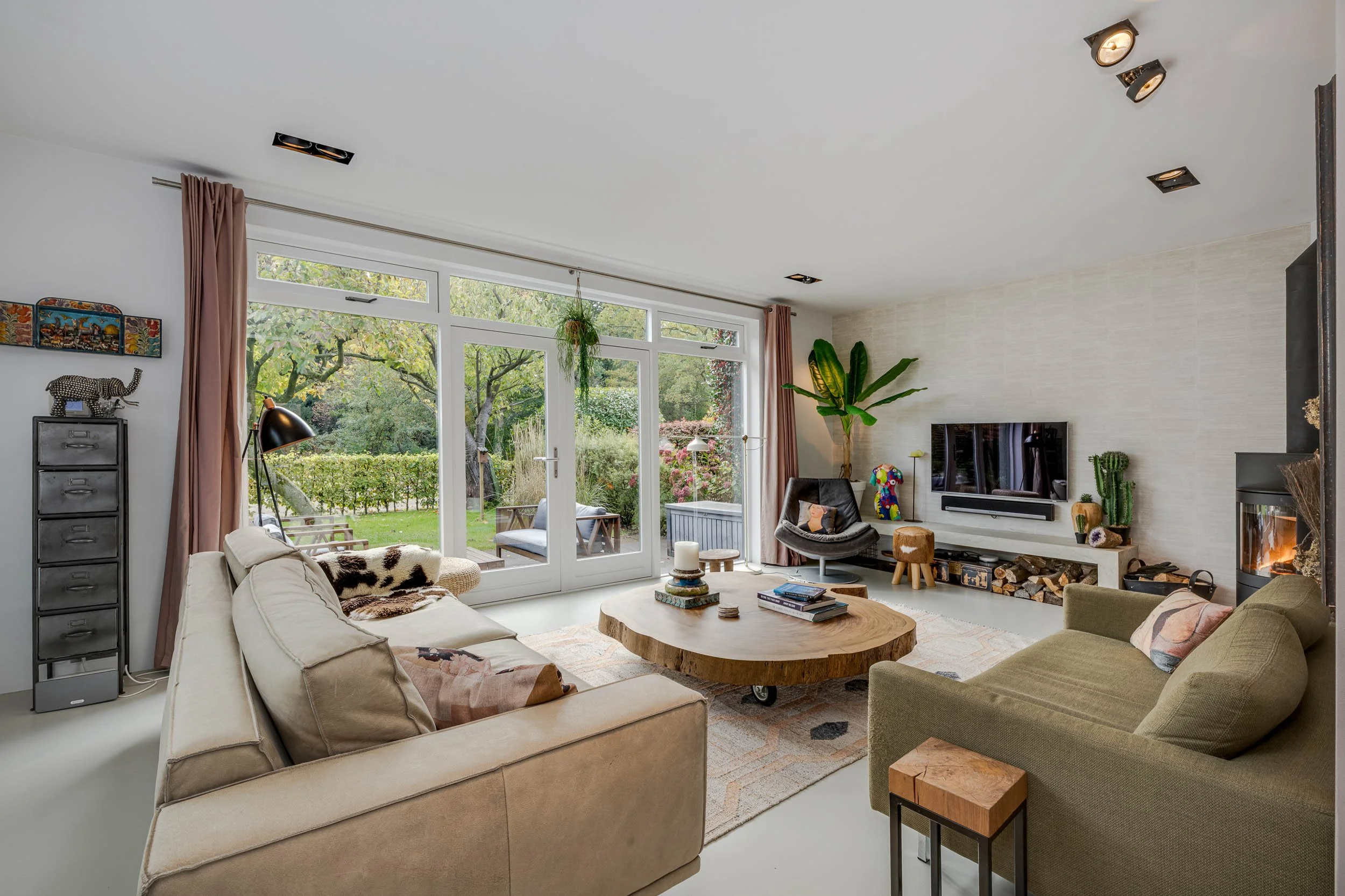 Woonkamer met banken, tv, houtkachel, grote ramen met uitzicht op tuin, planten, en decoraties.