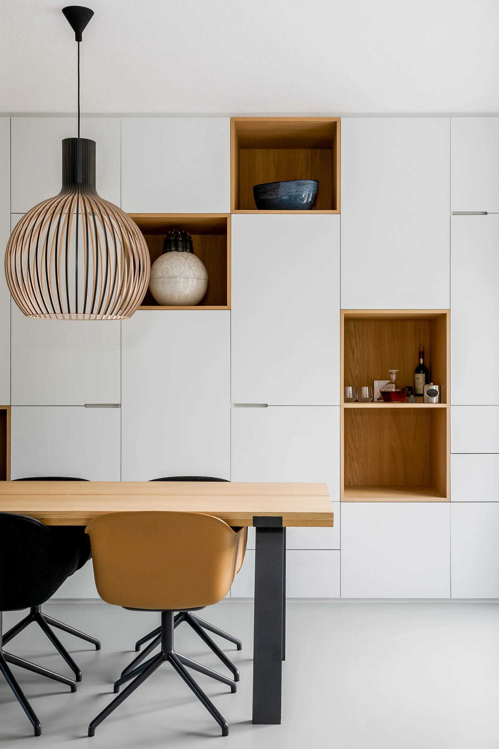 Moderne eetkamer met witte kasten, open vakken met decoratieve spullen, hanglamp en beige en zwarte stoelen rondom een houten tafel.