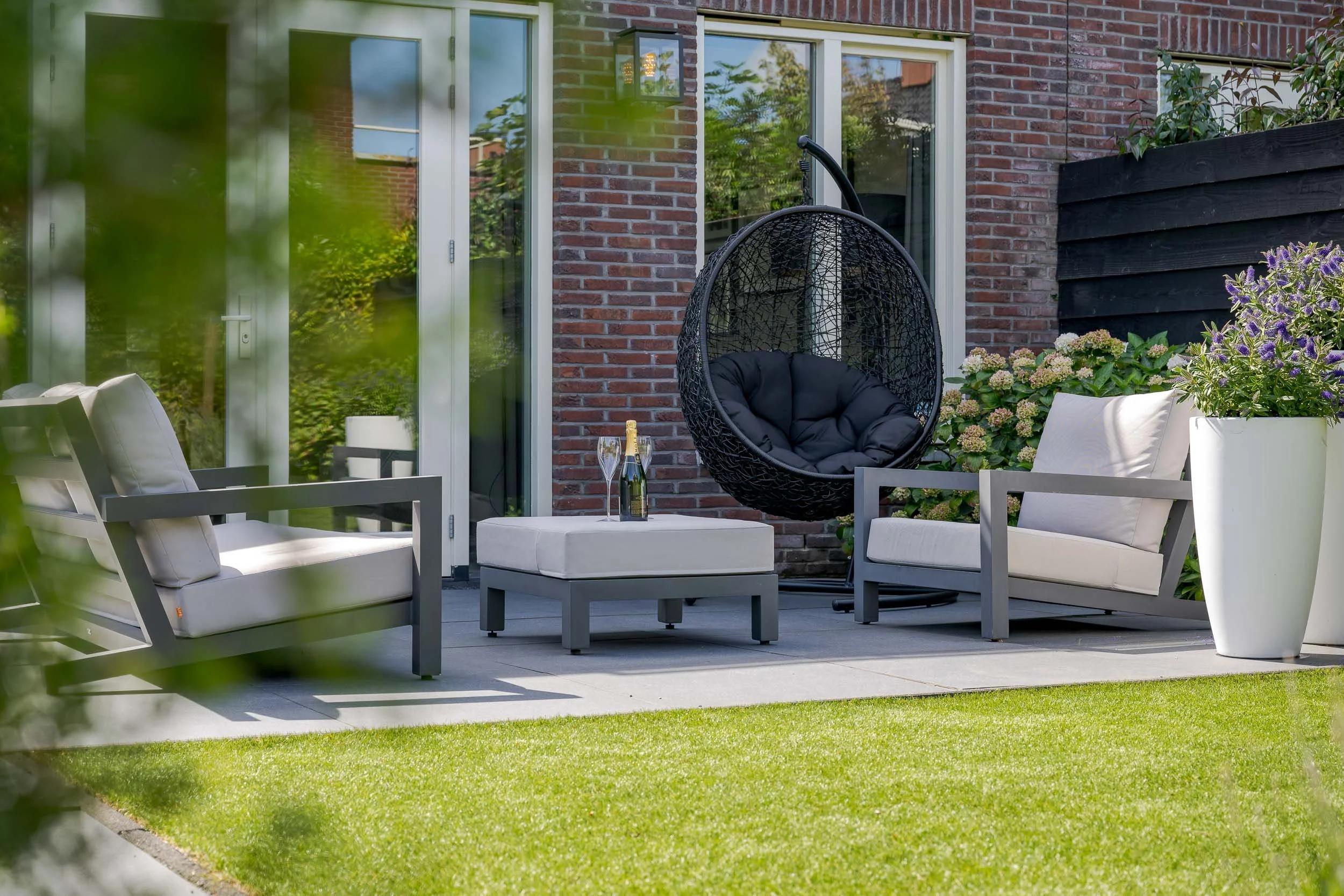 Een buiten terras met witte lounge stoelen, een kleine tafel met een fles en twee glazen, een hangstoel met kussen, en grote planten in witte potten, gelegen voor een bakstenen huis met grote ramen en een deur.