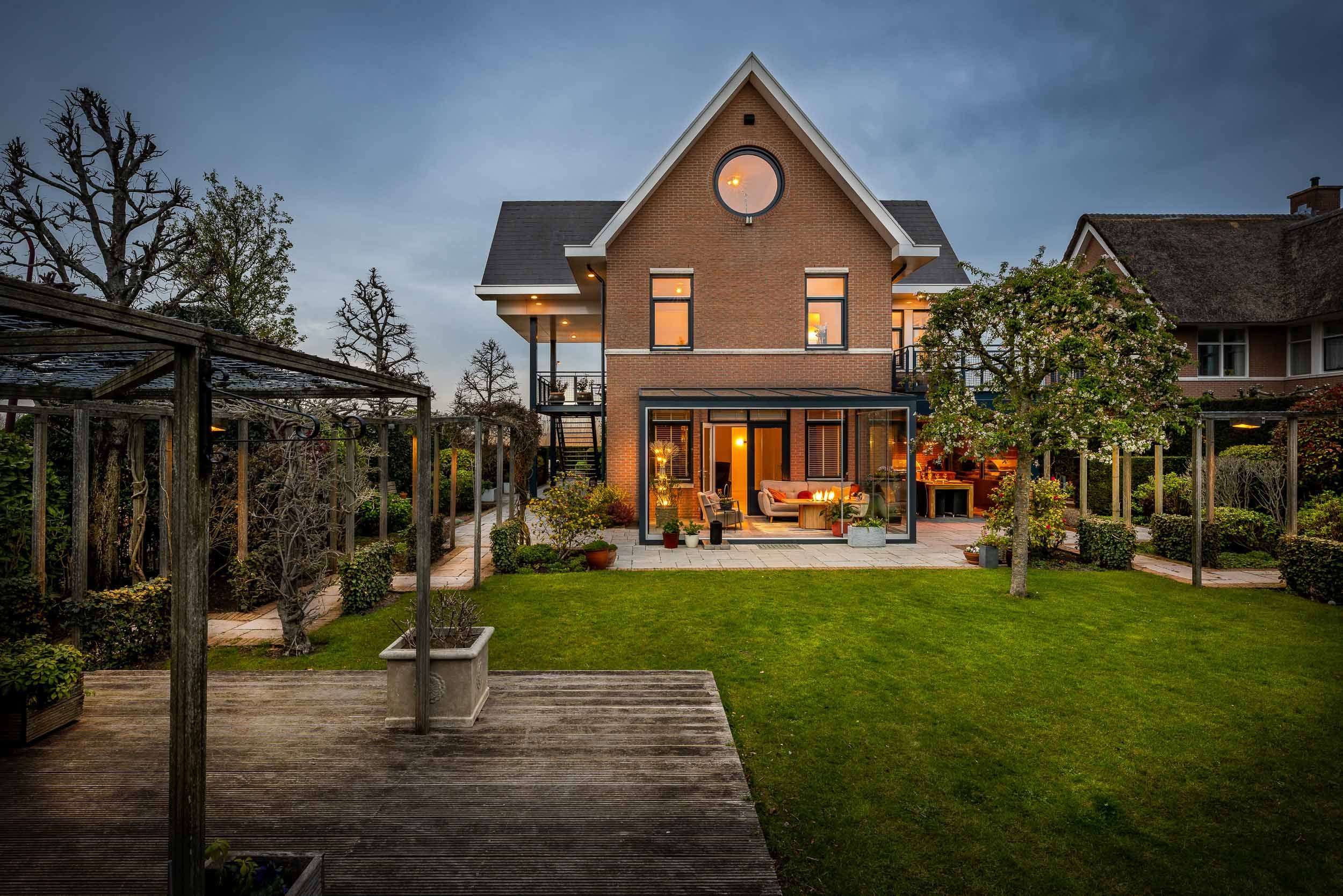 Een modern huis met een tuin bij schemering, verlichte binnenverlichting, een terras met meubels en tuinplanten, omgeven door een grasveld en bomen.
