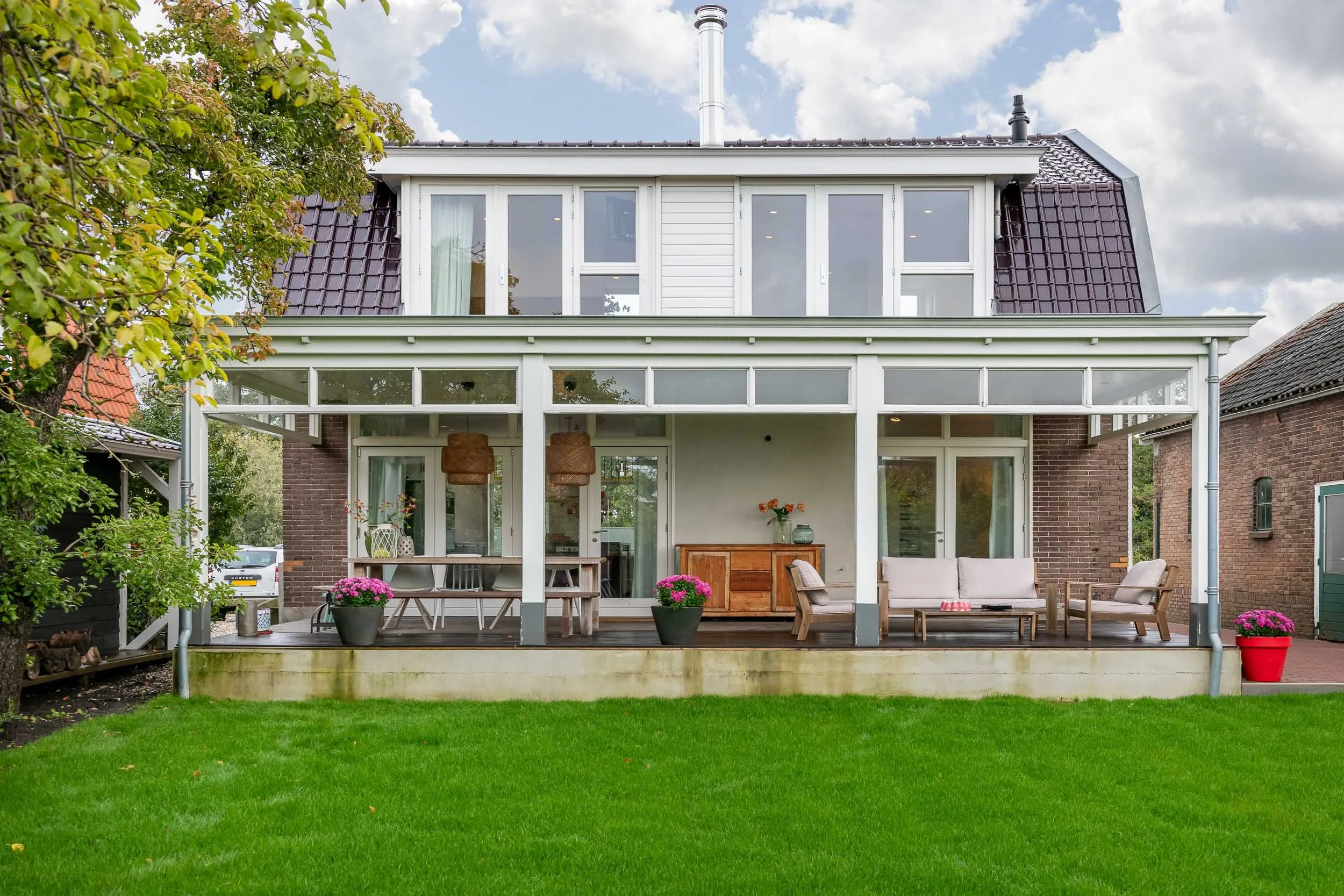 A modern house met een grote terras met tuinmeubilair en bloemen, omgeven door groen gras en bomen onder een bewolkte hemel.
