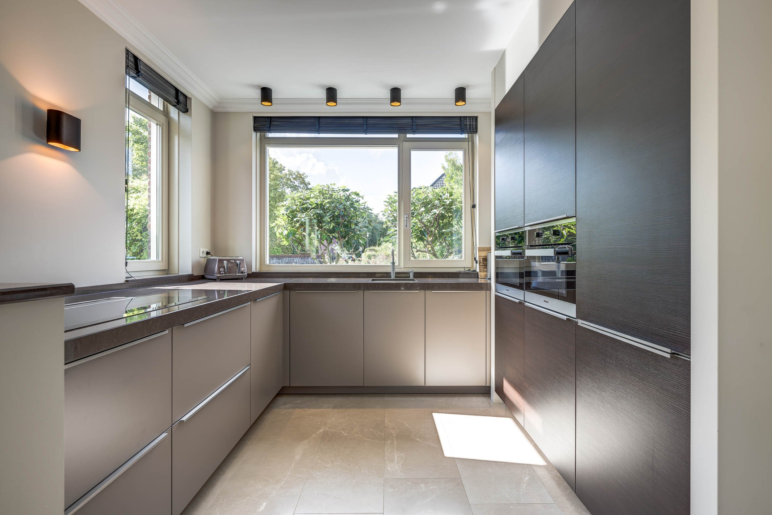 Moderne keuken met grote ramen, beige kasten, ingebouwde oven en koelkast, zwart plafondlampen, en uitzicht op groene bomen buiten.