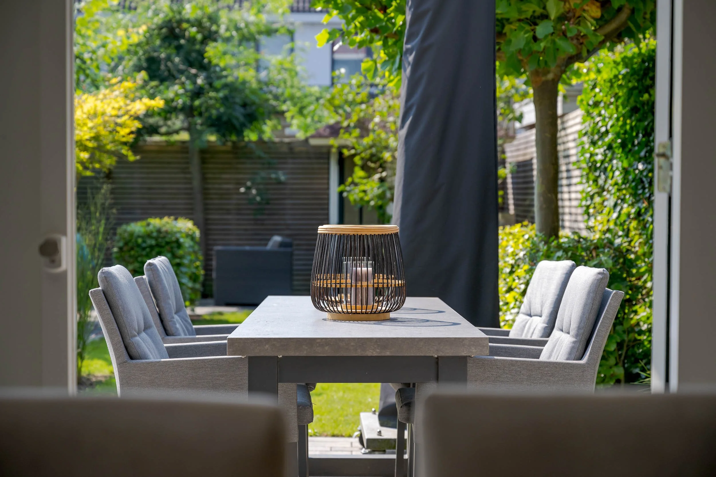 Gezicht op een tuin met een eettafel en stoelen, gekaderd door een open deur, met groene bomen en een houten schutting op de achtergrond.