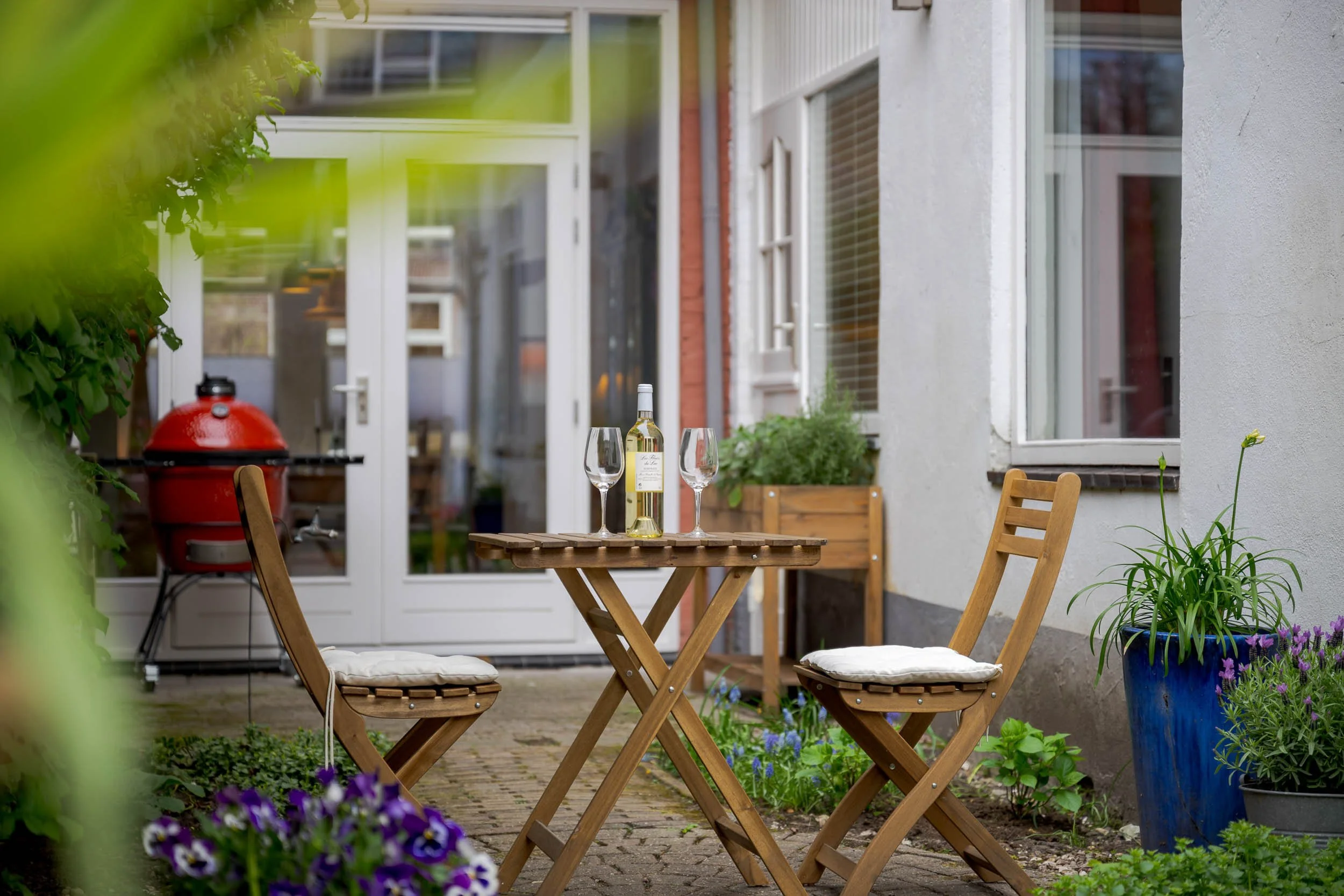 Een tuin met een houten tafel en stoelen, een fles witte wijn en twee wijnglazen, omringd door bloemen en planten, met een huis op de achtergrond.