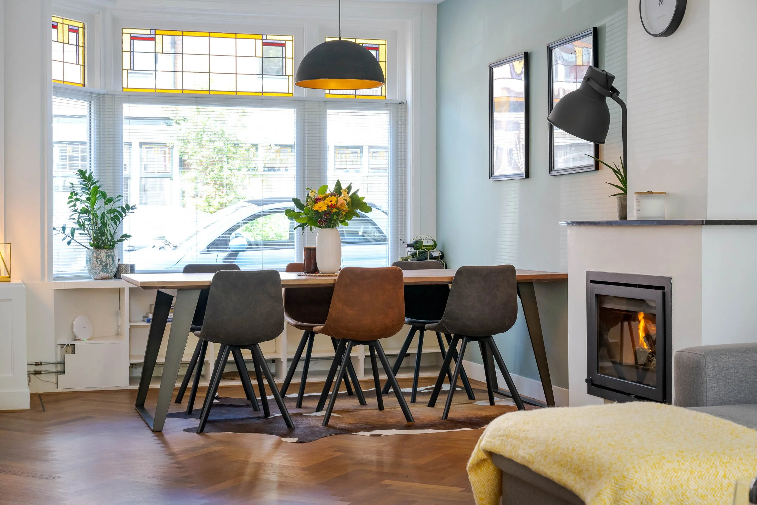 Een knusse woonkamer met een grote eettafel, omringd door verschillende stoelen, een plantenbak, een bloemenvaas, en een open haard met een vuur. Grote ramen met jaloezieën en sfeervolle verlichting zorgen voor een gezellige ambiance.