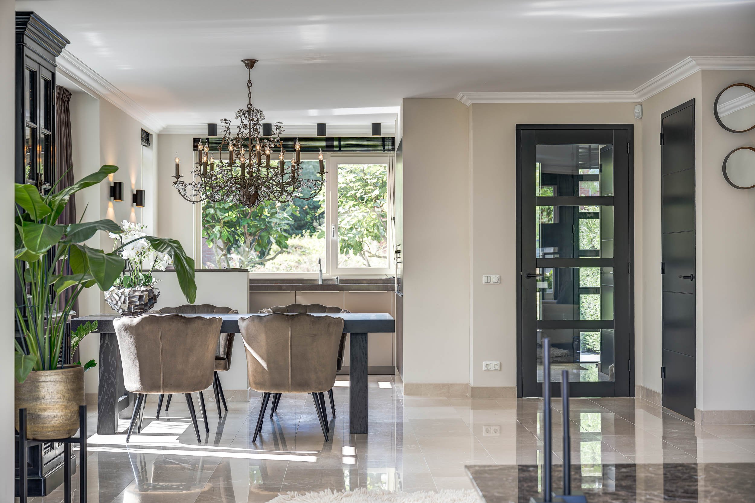 Moderne woonkeuken met grote ramen, zwarte deur, chandelier, planten en zitplaatsen.