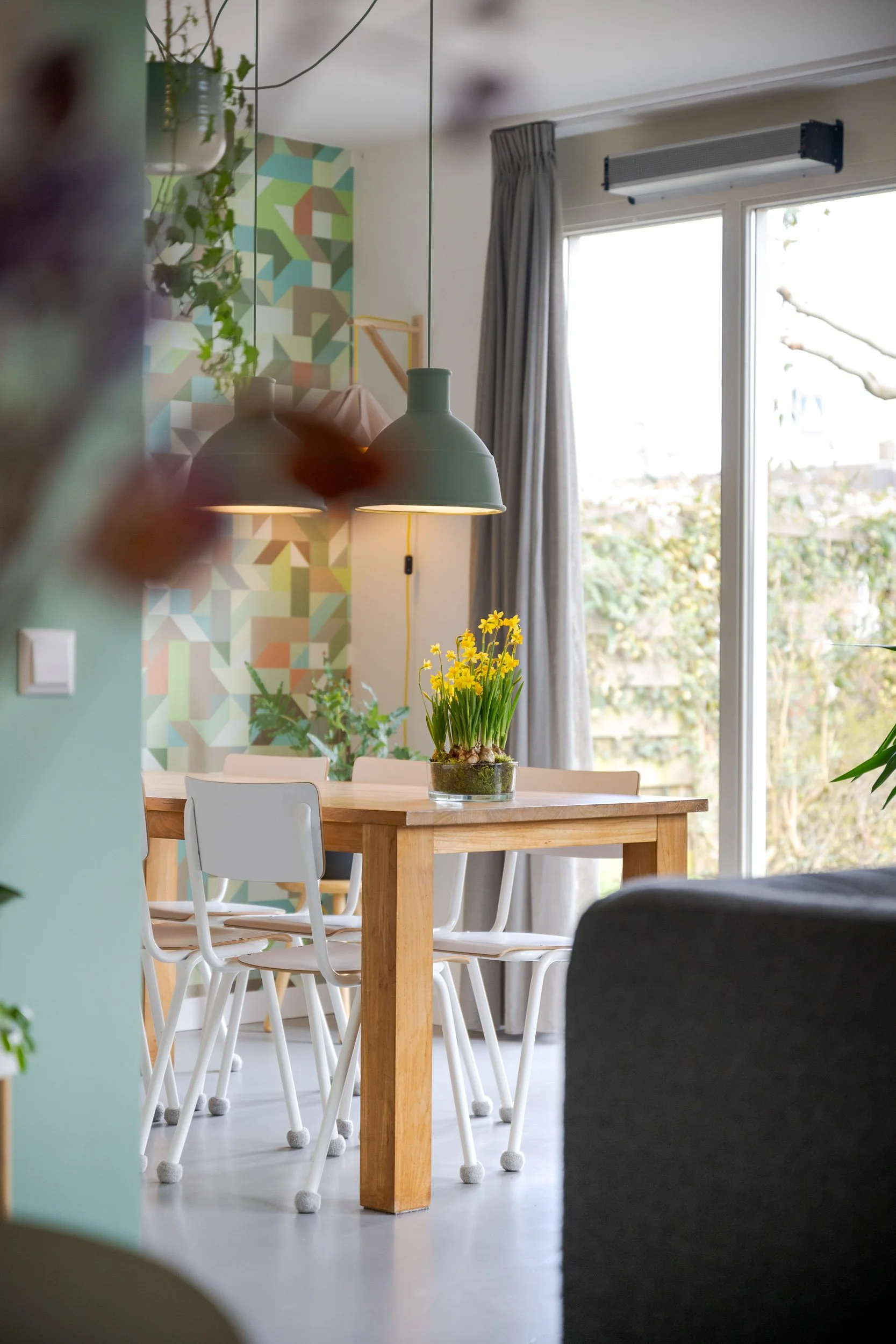 Een eetkamer met een houten tafel, witte stoelen en een plant met gele bloemen op tafel. In de achtergrond een muur met een geometrisch patroon en een raam met grijze gordijnen.