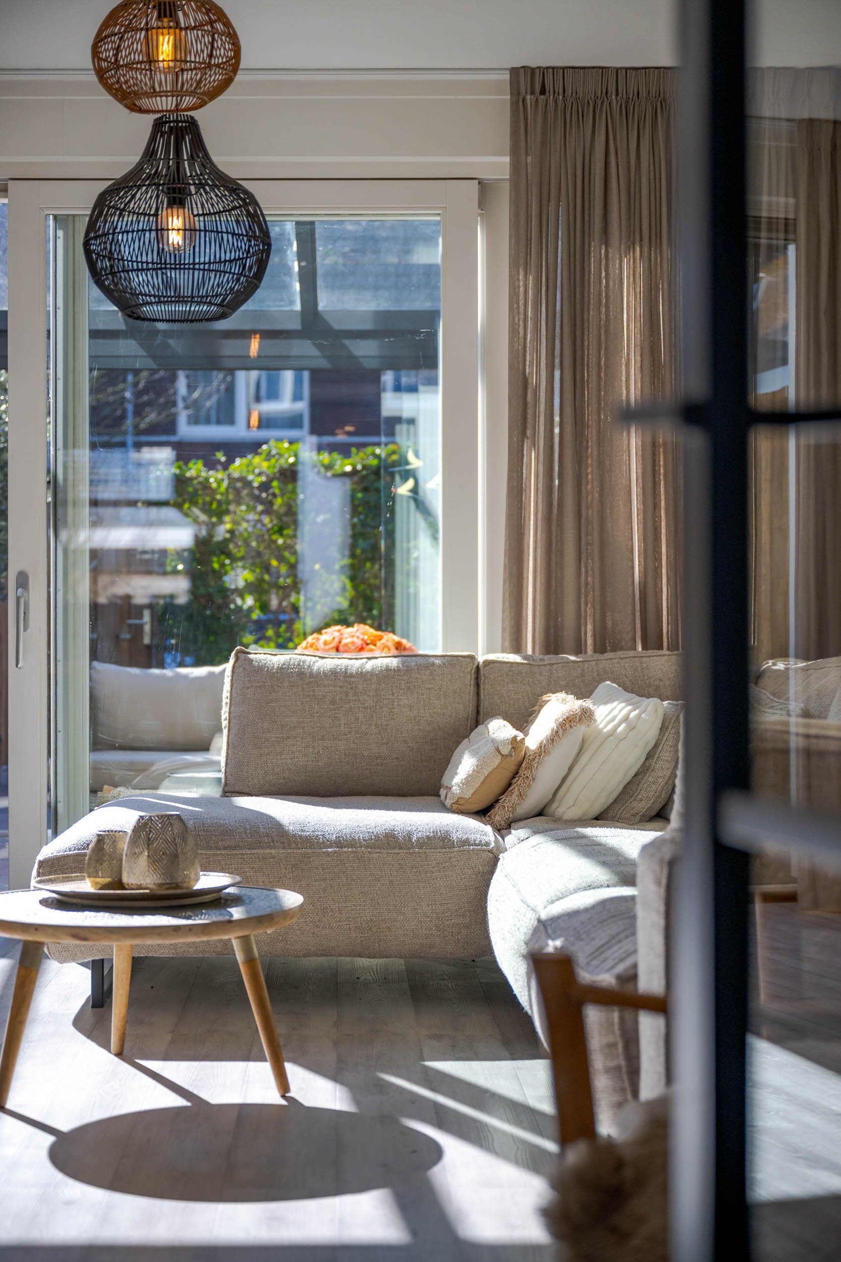 Een woonkamer met een beige bank, beige en crème kussens, een ronde houten salontafel met decoratieve voorwerpen, grote ramen met beige gordijnen, een dubbele hanglamp met ronde rieten kappen, en een terras met tuin en huizen buiten.