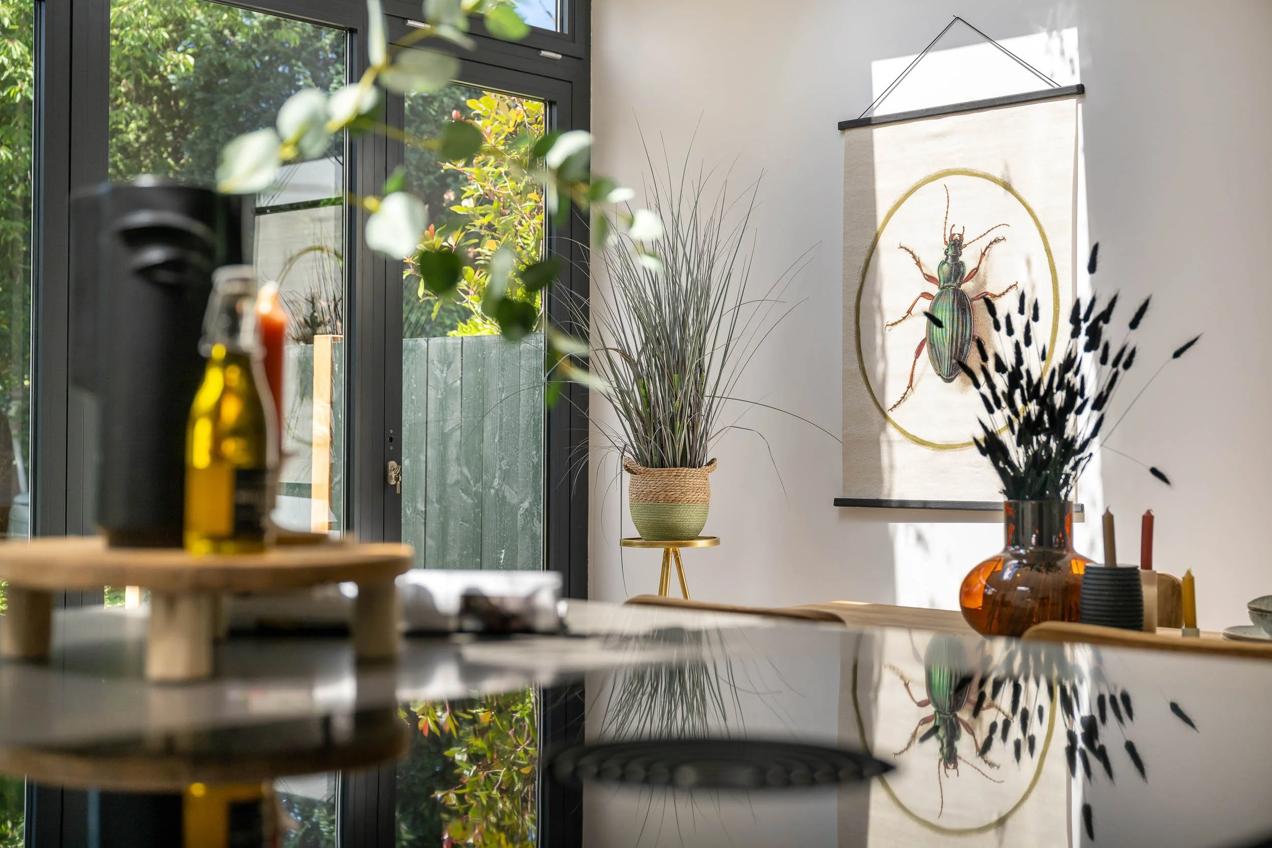 Interieur van een modern huis met grote ramen, een muurdecoratie van een kever, en verschillende planten en decoratieve voorwerpen op een tafel.