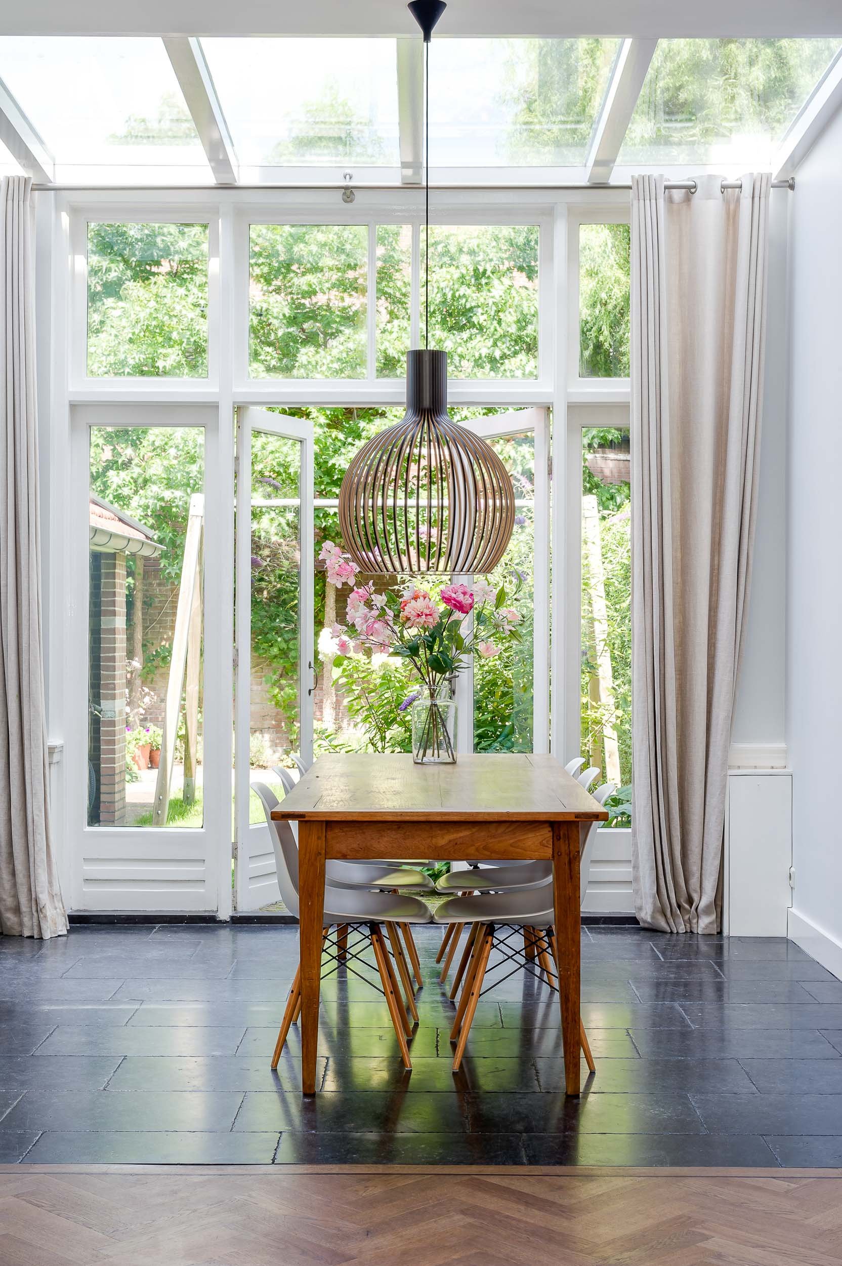 Een eettafel met vier stoelen voor grote ramen met uitzicht op een tuin, een hanglamp boven de tafel, en een vaas met roze bloemen.