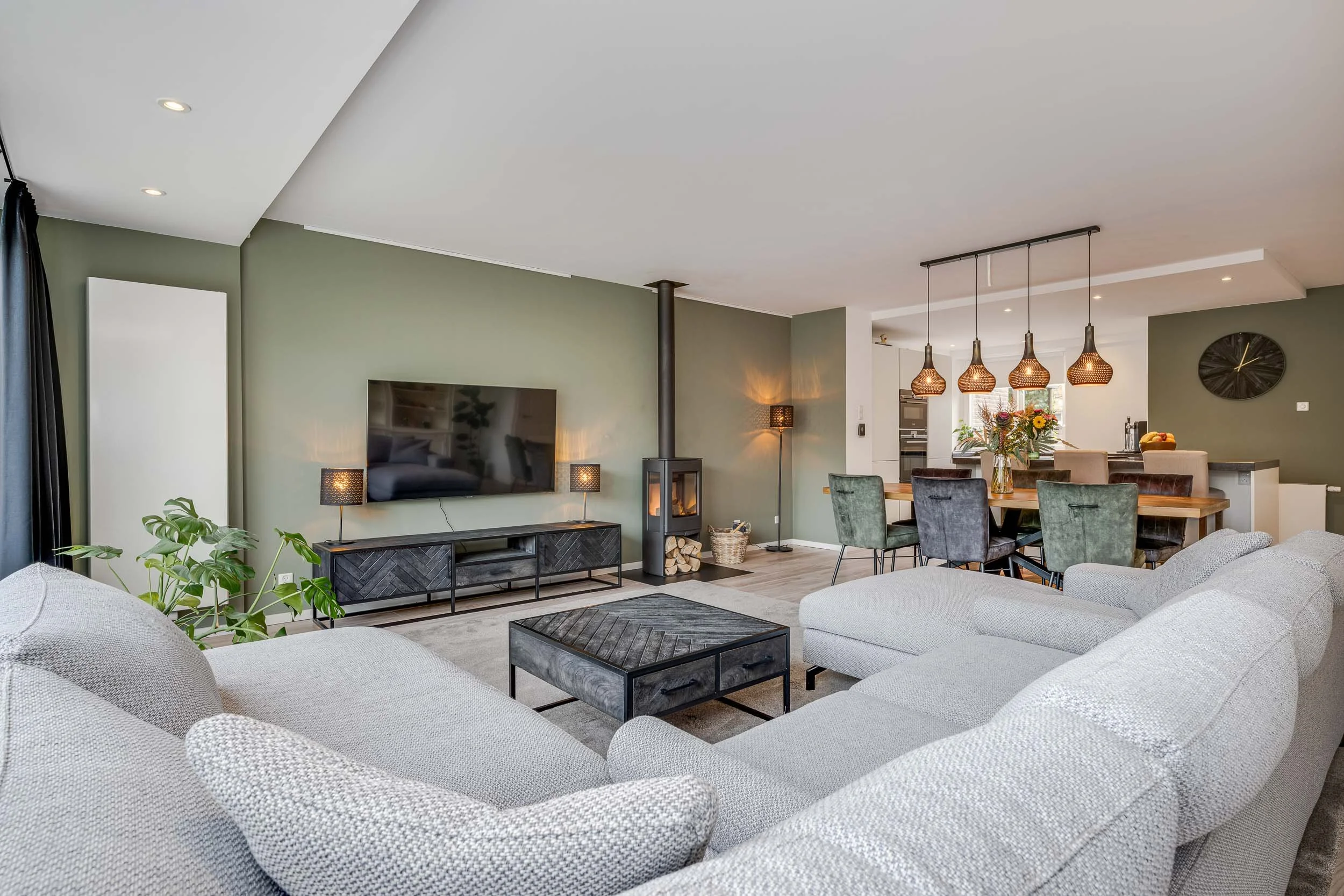 Een moderne woonkamer met een grote witte bank, een tv aan de muur, een houtkachel, planten en een eettafel met stoelen onder plafondlampen.
