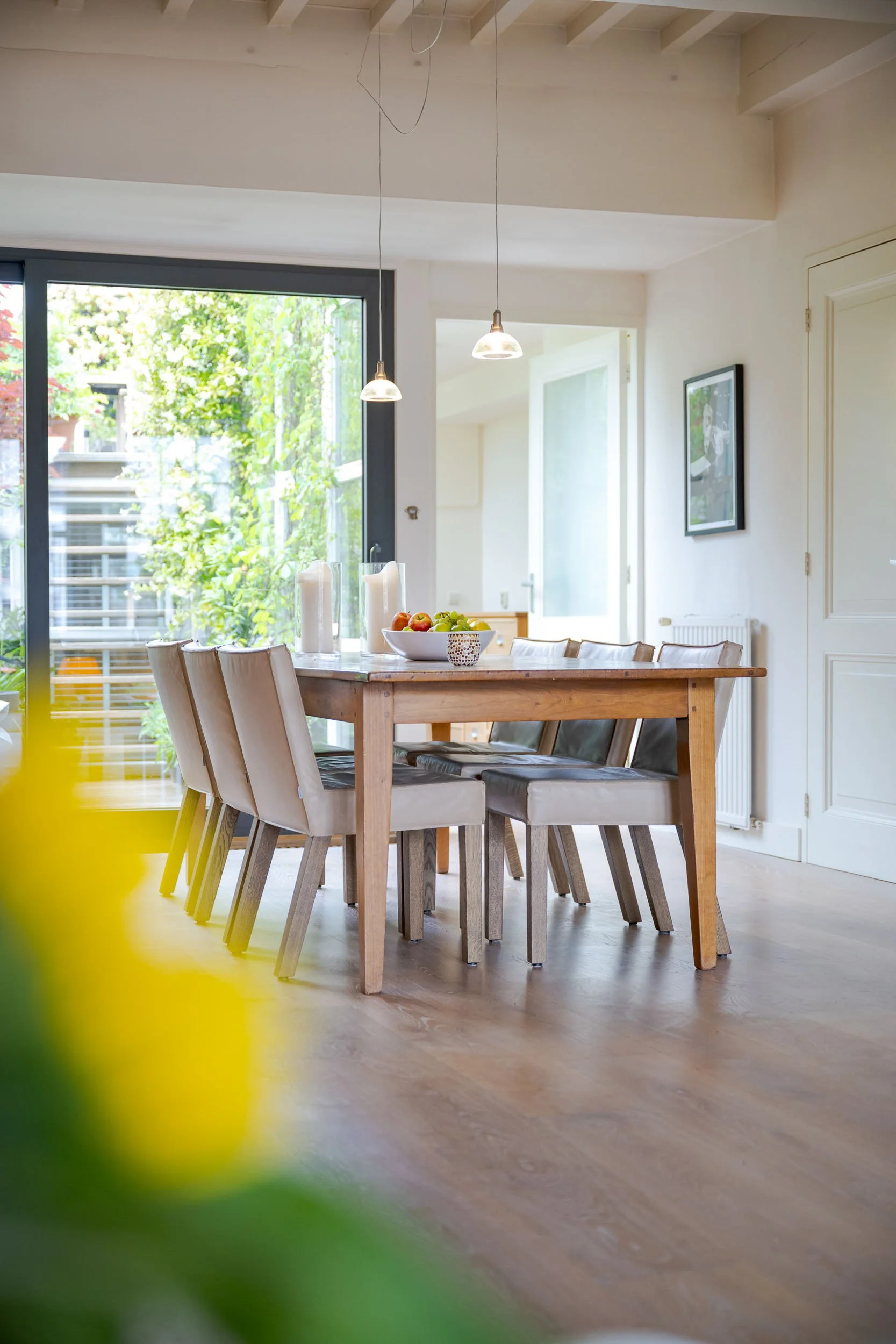 Binnenkant van een lichte, moderne eetkamer met een houten tafel, beige stoelen, een fruitschaal met appels, kaarsen en één hangende lamp, grote glazen deuren die naar een tuin leiden, en een foto aan de muur.
