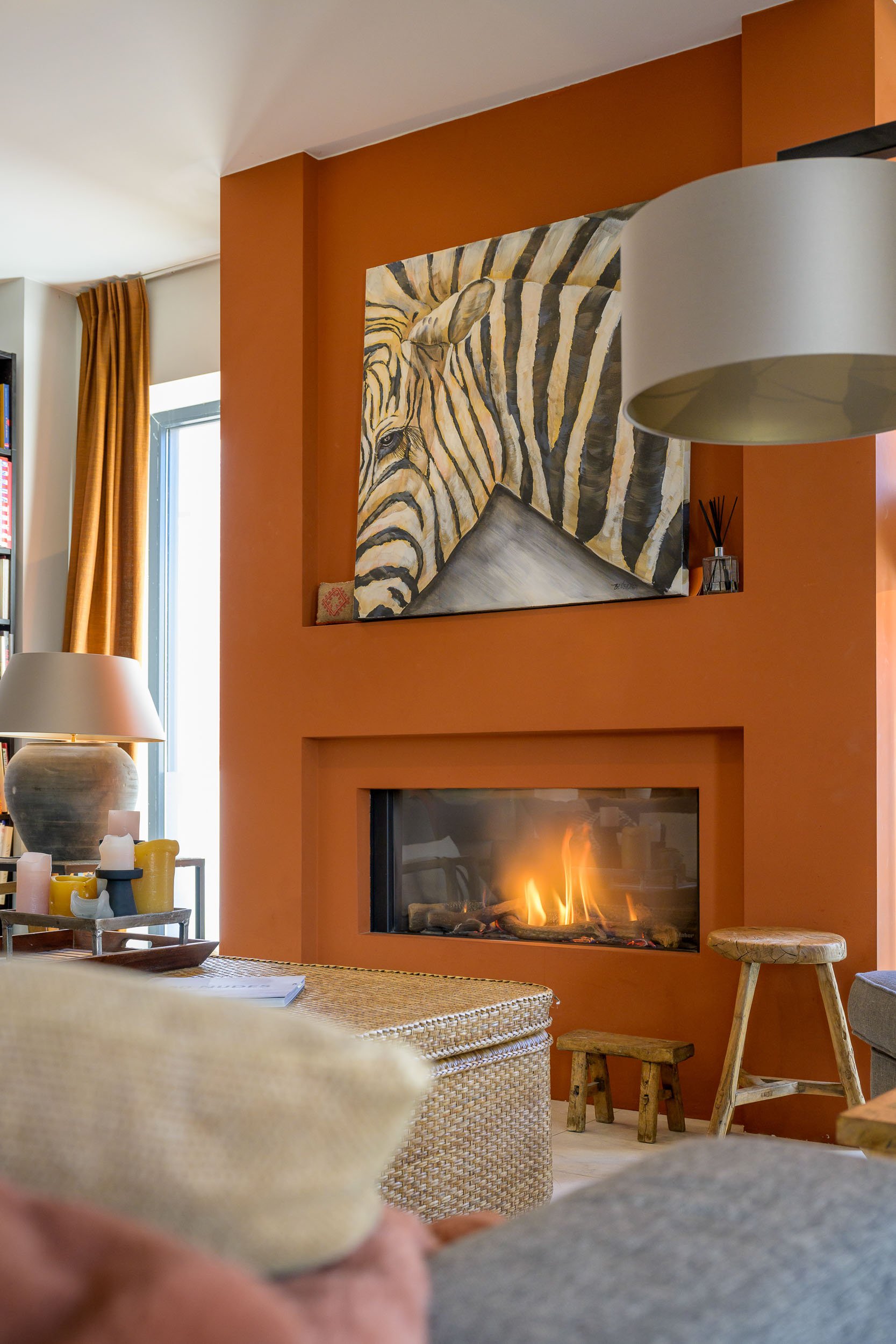 Een woonkamer met een haardvuur, een schilderij van een zebra aan de muur, een witte lamp, een tafel met kaarsen en een klein krukje.