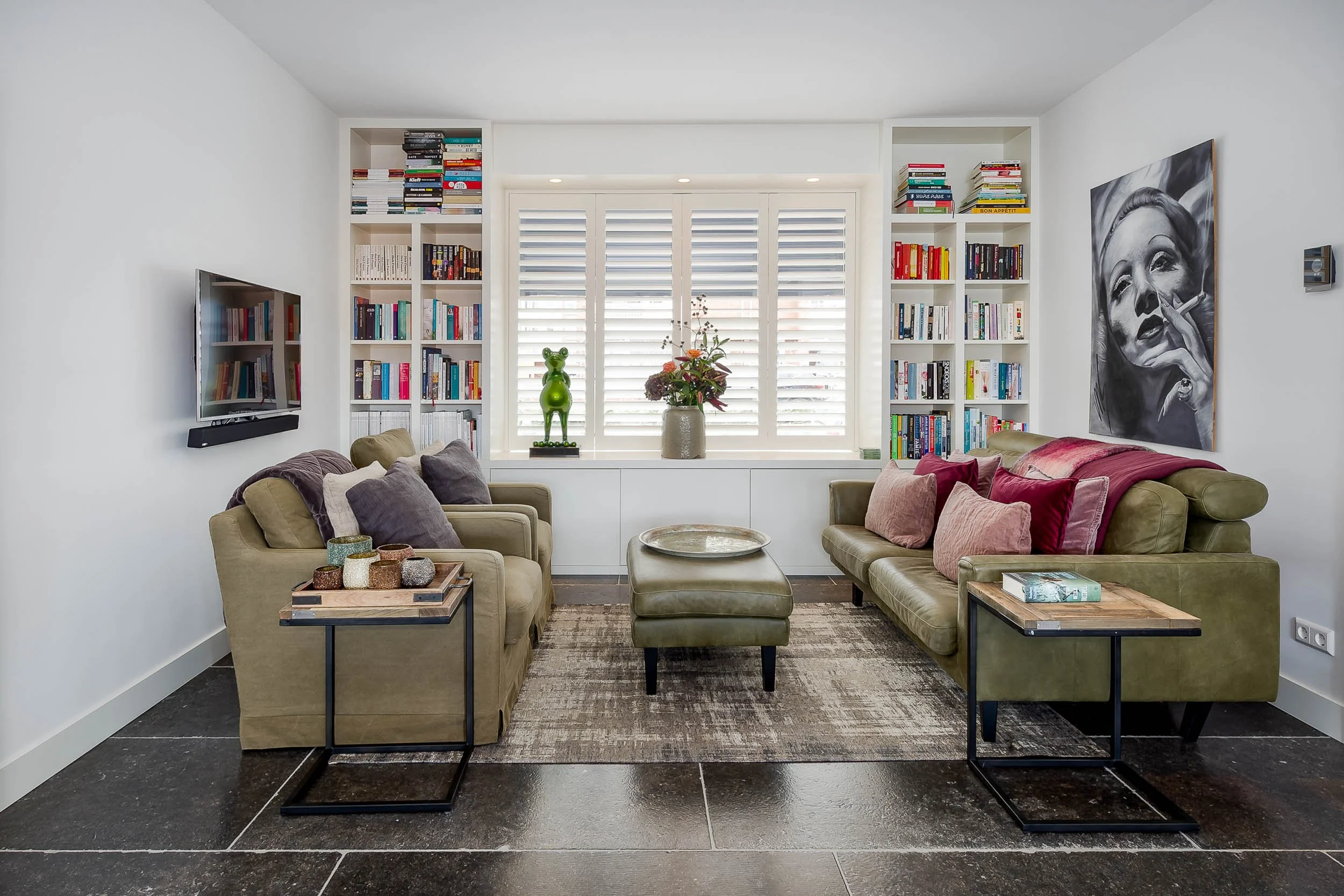 Een woonkamer met twee groene leren banken en een voetenbank, een salontafel met decoratieve vazen, ingebouwde boekenplanken, een grote venster met witte shutters, bloemen in een pot en een zwart-wit schilderij aan de muur.