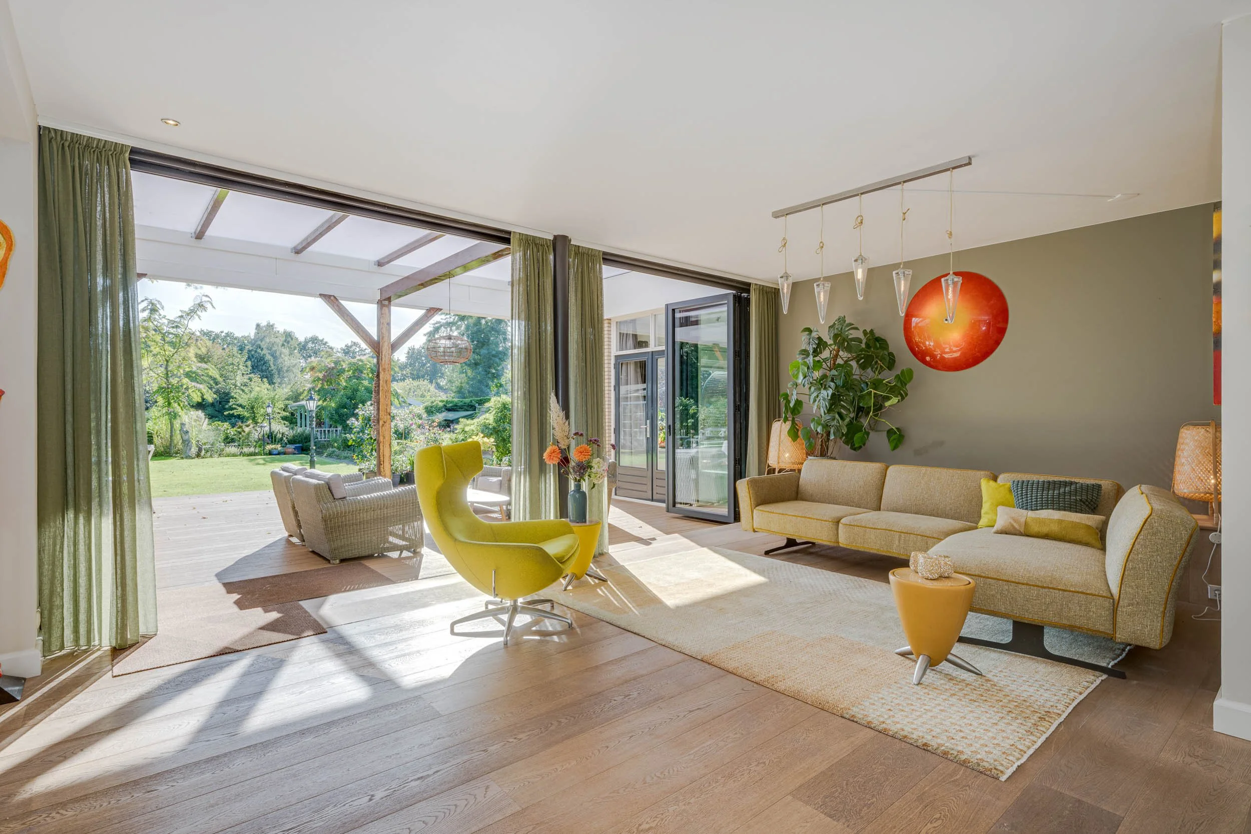 Een lichte woonkamer met grote ramen en openslaande deuren die leiden naar een tuin. Binnen staan gele en groenige stoelen, een roomkleurige bank en een lage tafel met decoraties. Er hangt een moderne hanglamp met glazen pepers en er is een groene pl