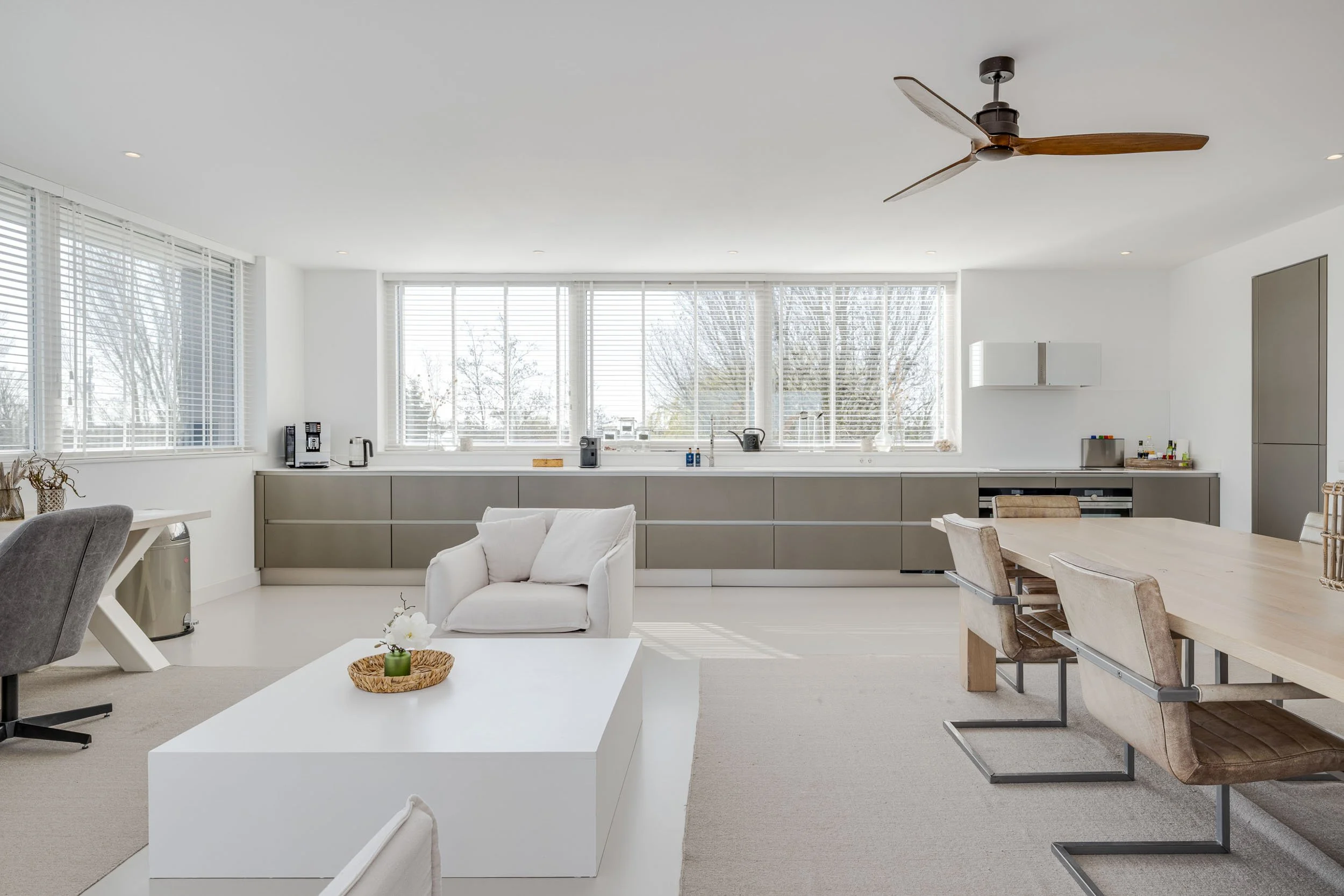 Modern open keuken en eetkamer met grote ramen en witte jaloezieën, lichte kleuren, houten details, en een plafondventilator.