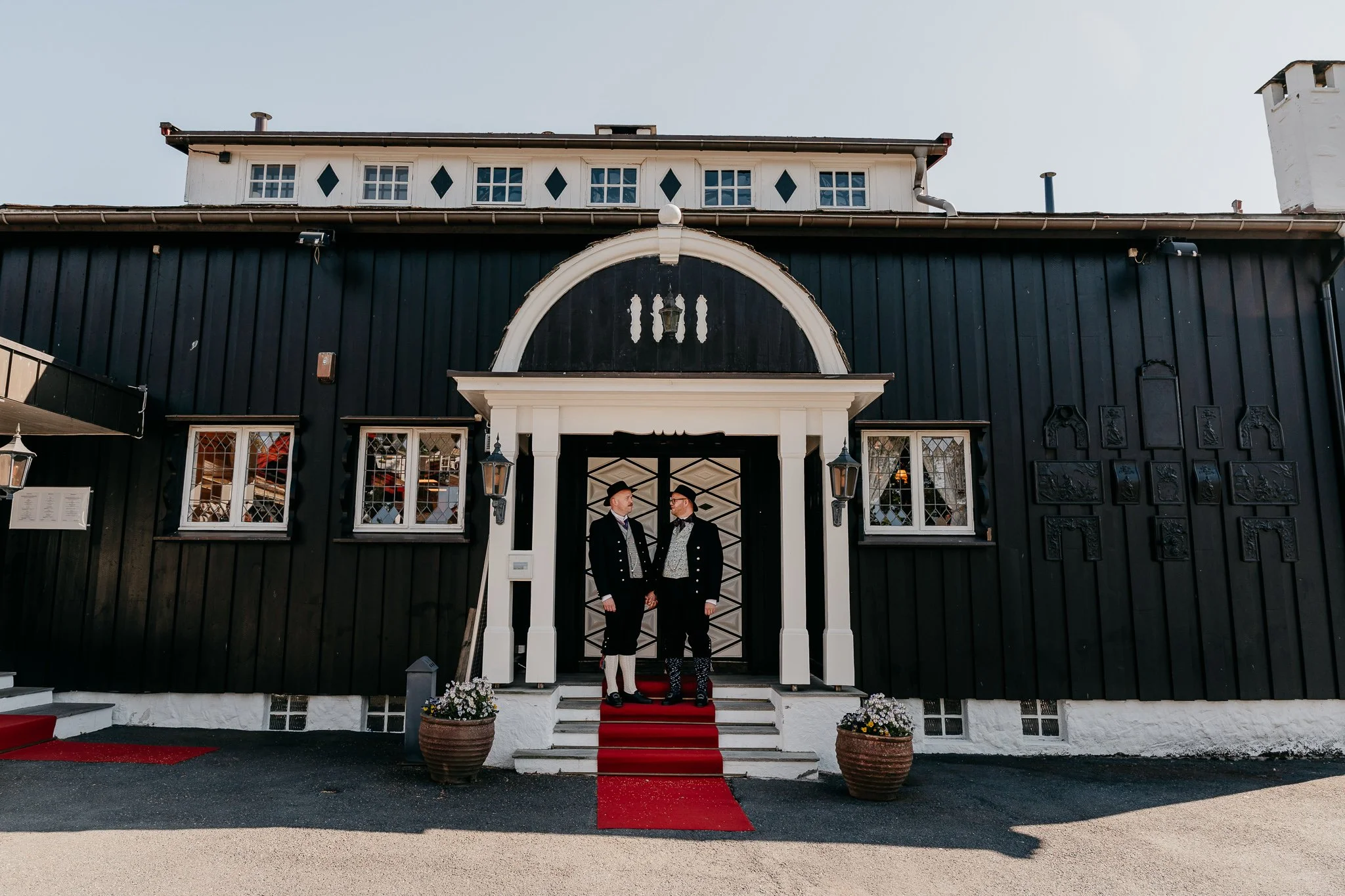 To menn i bunad holder hender utenfor et ærverdig bygg.