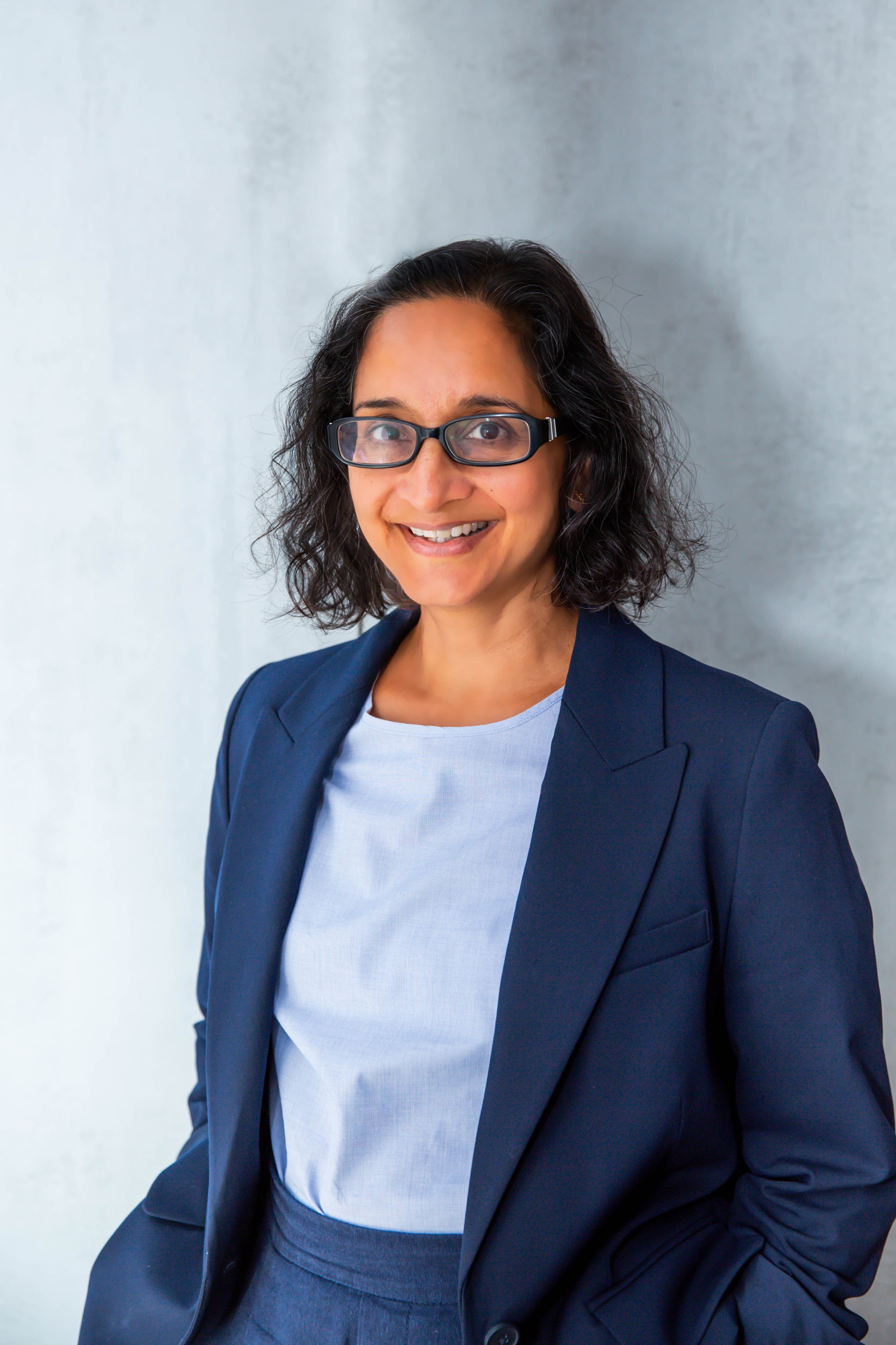 A woman with curly dark hair and glasses, smiling, wearing a navy blazer and white shirt, standing against a plain light gray background.