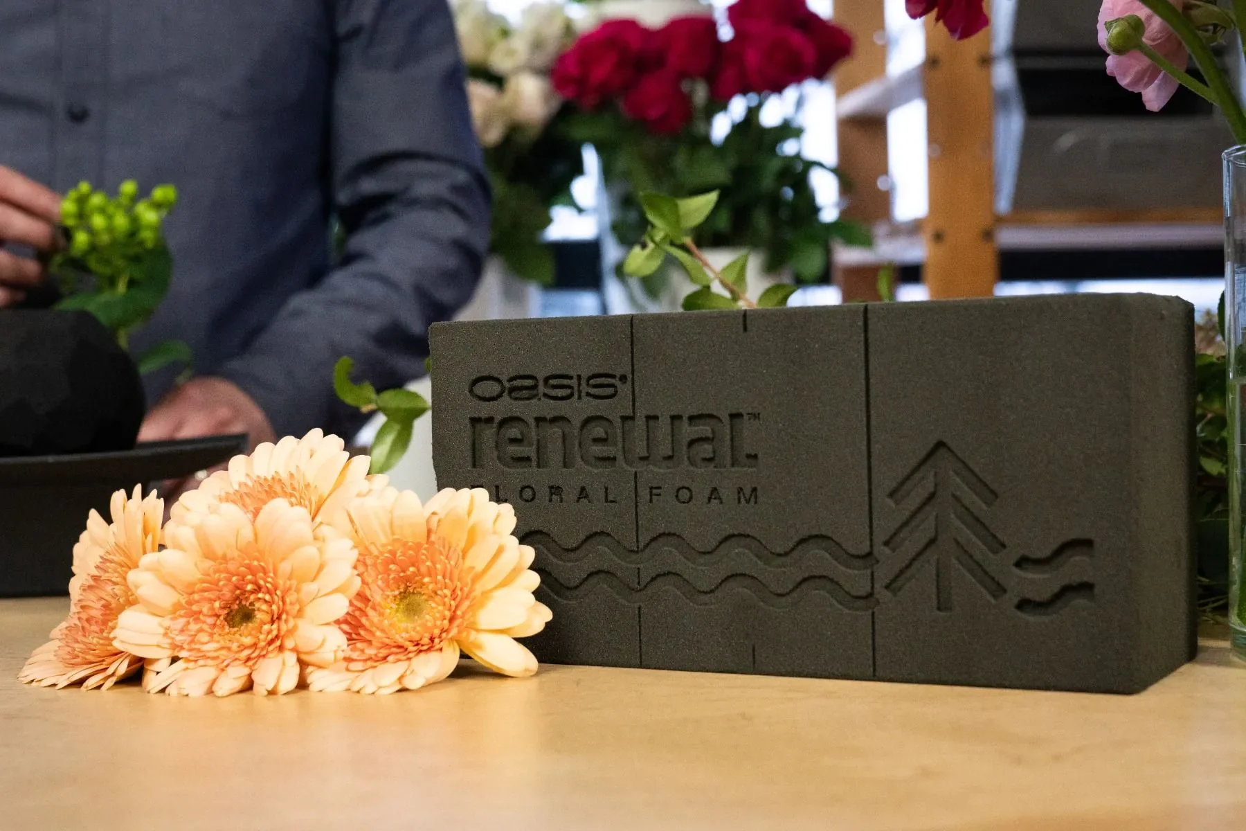 A floral foam brick labeled 'Oasis Natural Floral Foam' is on a wooden table, with peach-colored flowers and some green leaves in front of it. In the background, there are various flowers and a person in a gray shirt.