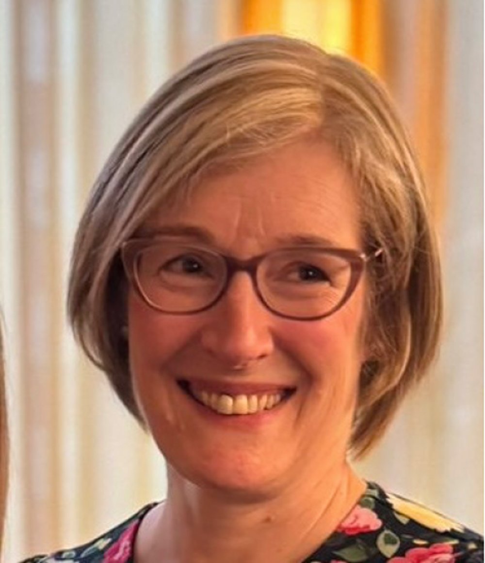 Black and white photo of an older woman wearing glasses, a patterned scarf, and a leopard print top, standing in front of a plain wall.