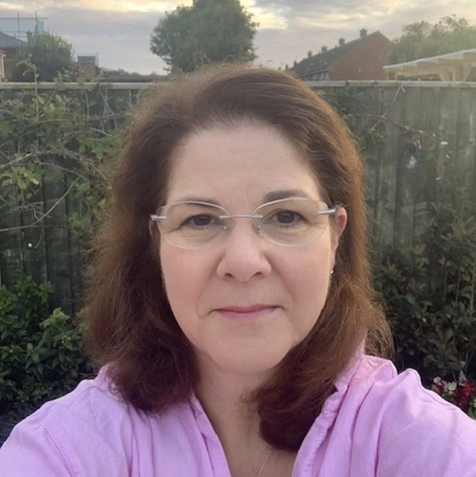 A woman with shoulder-length dark hair, wearing glasses and a light-colored shirt, looking at the camera, outdoors with a fence, trees, and houses in the background.