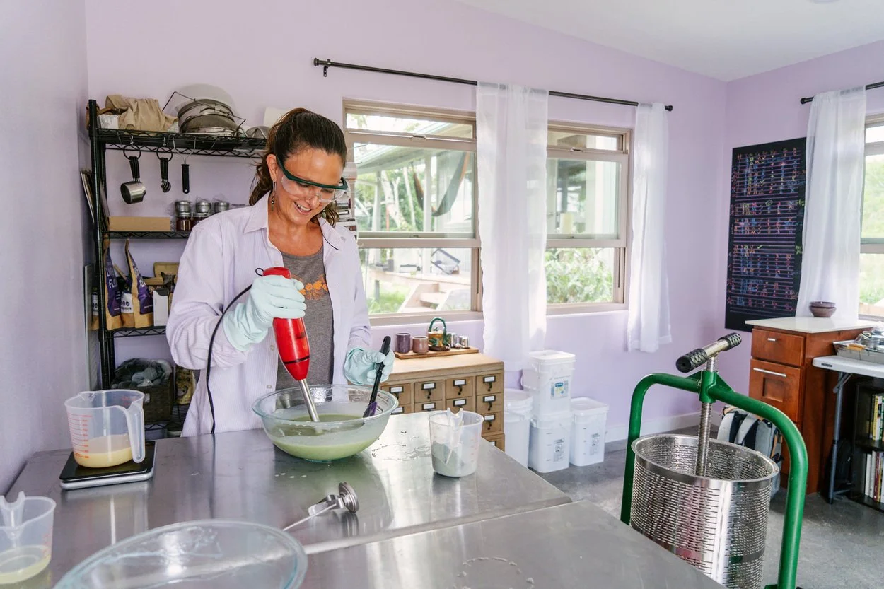 Woman wearing safety glasses and gloves blending a green liquid in a bowl with a red immersion blender in a light purple room kitchen.