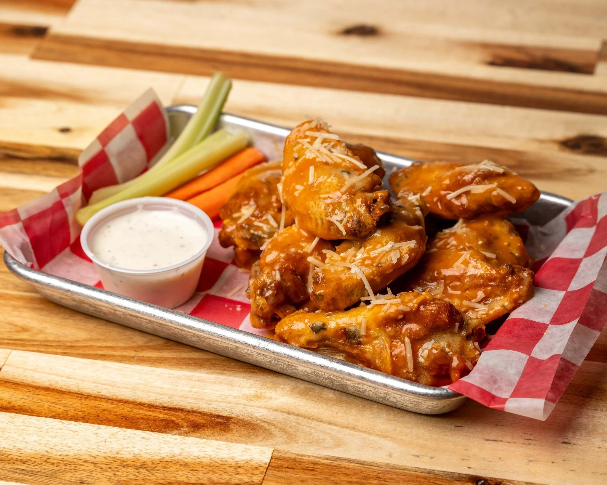 A tray of sauced chicken wings with ranch dressing, celery and carrot sticks on a wooden table.