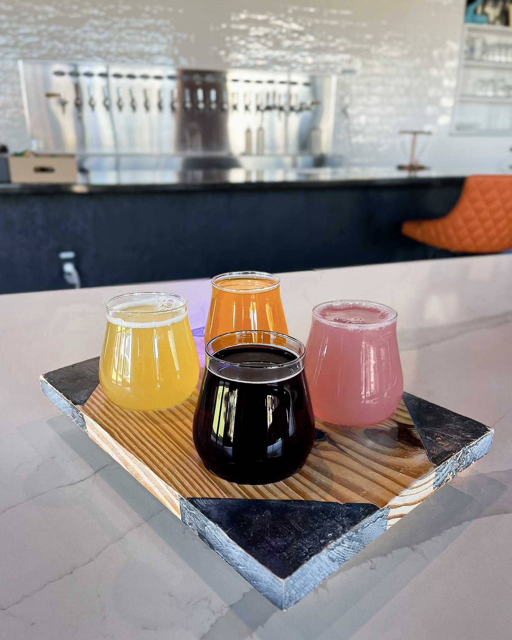 Four tasting glasses of golden, orange, dark and pink beer on a wooden flight board, with the taproom bar behind them.