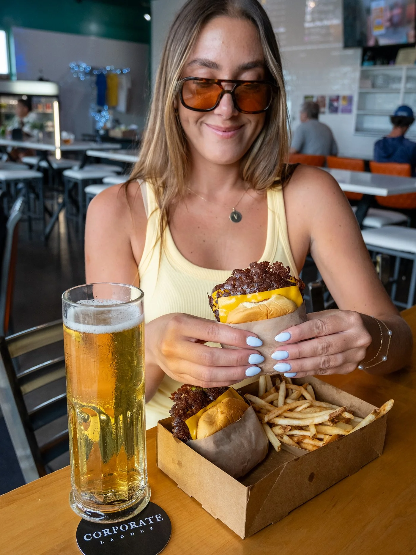 A taproom guest holding a cheeseburger beside a glass of golden beer and a tray of fries at a communal table.