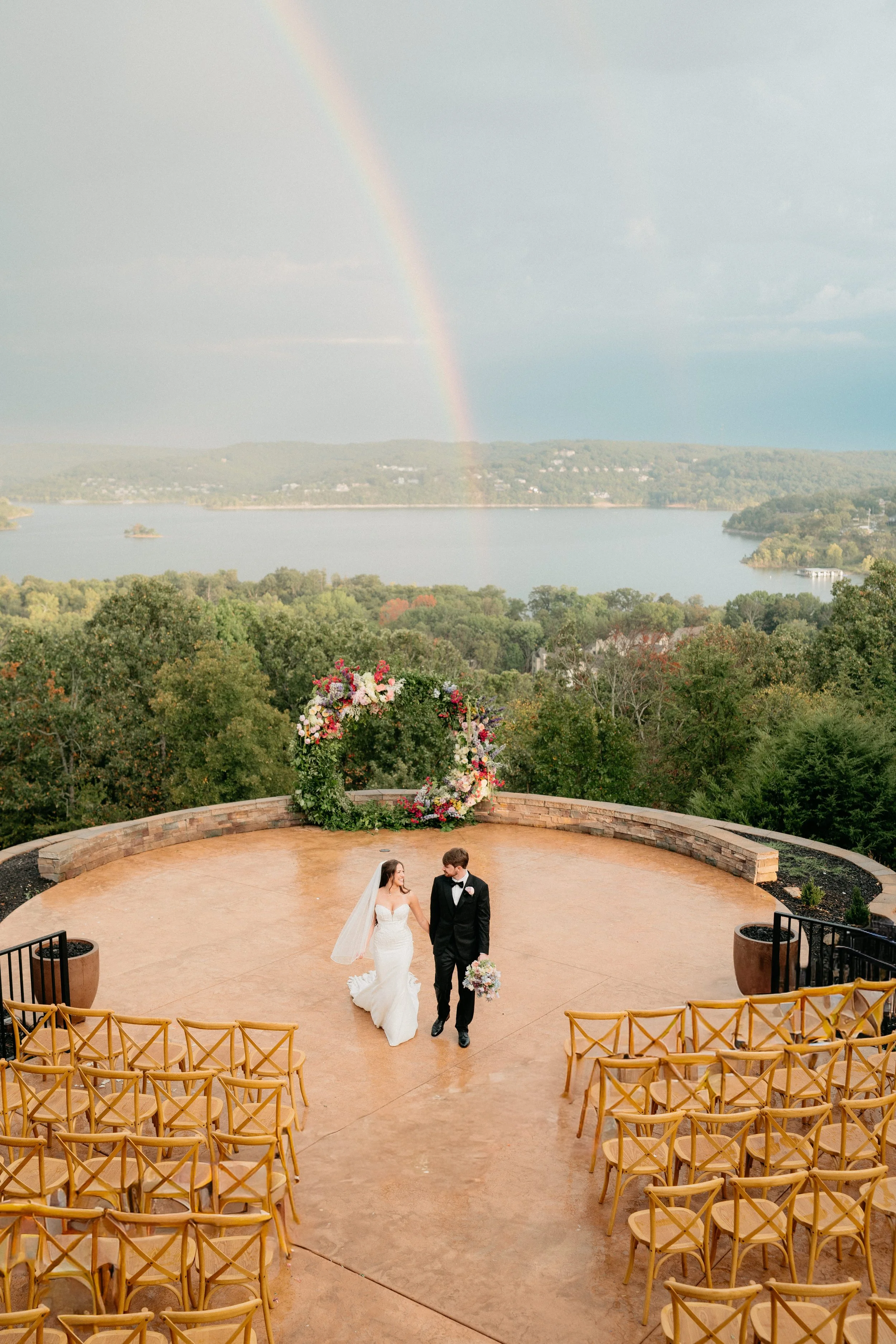 A newlywed couple holding hands walking on an outdoor wedding venue with a scenic view of a lake, hills, trees, and a rainbow in the sky.