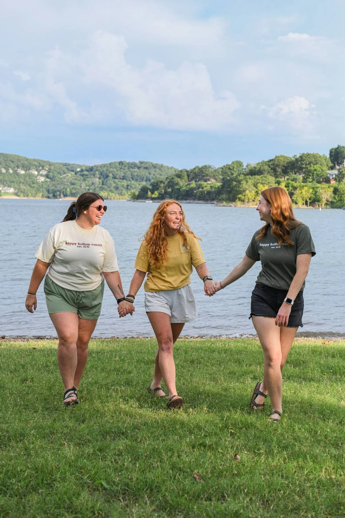 Three women holding hands and walking on grass near a lake, smiling and enjoying the outdoors.