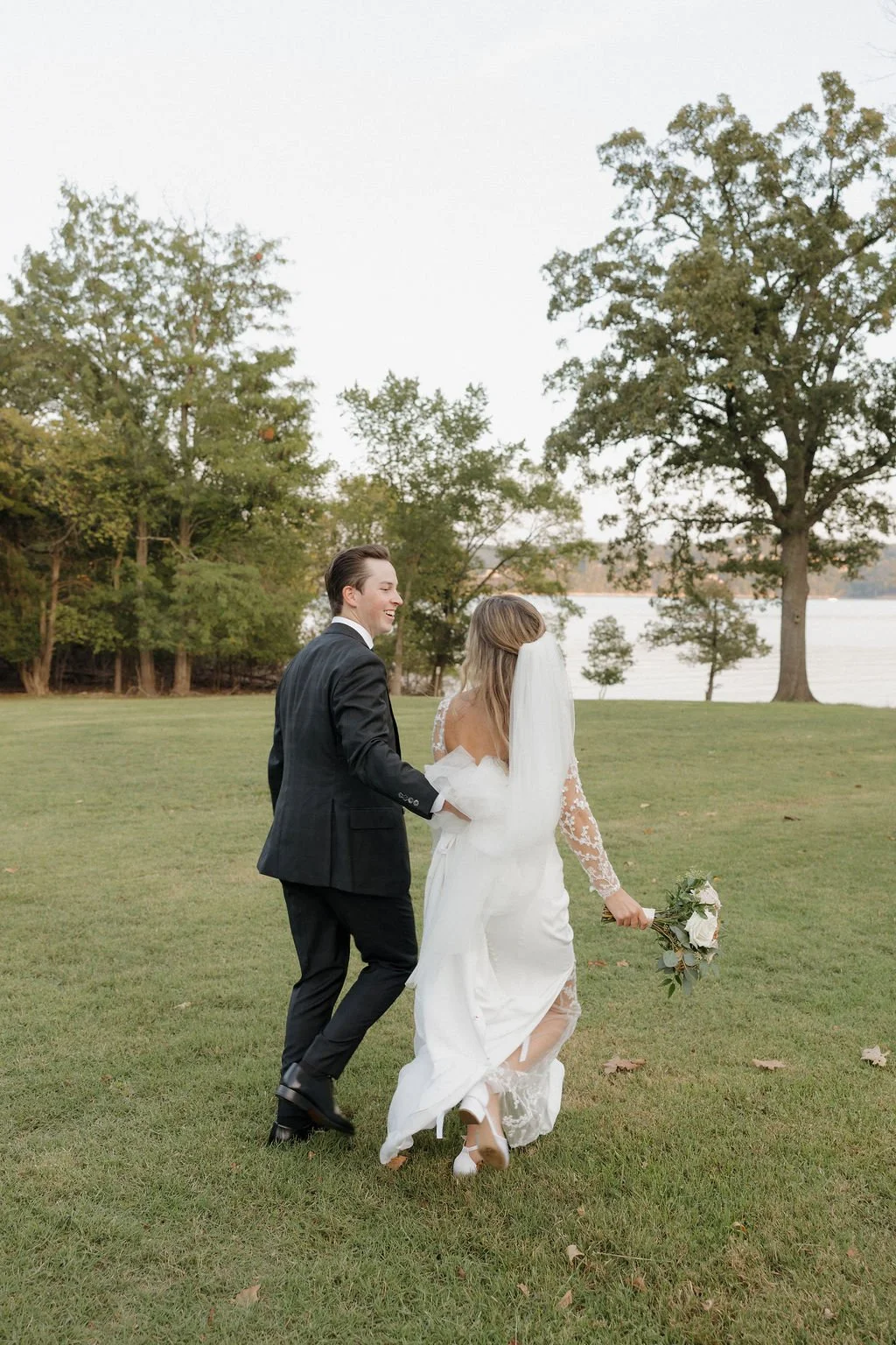 A newly married couple walking on grass near a lake in the Ozarks, holding hands and smiling, with trees and water in the background.