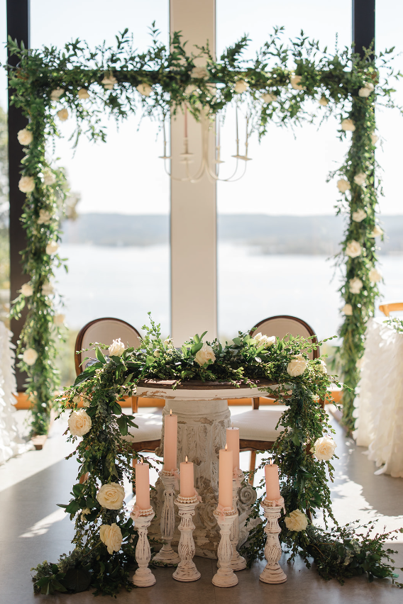 Elegant wedding altar with white roses and greenery, large windows overlooking a lake, decorated with a floral arch and candles.