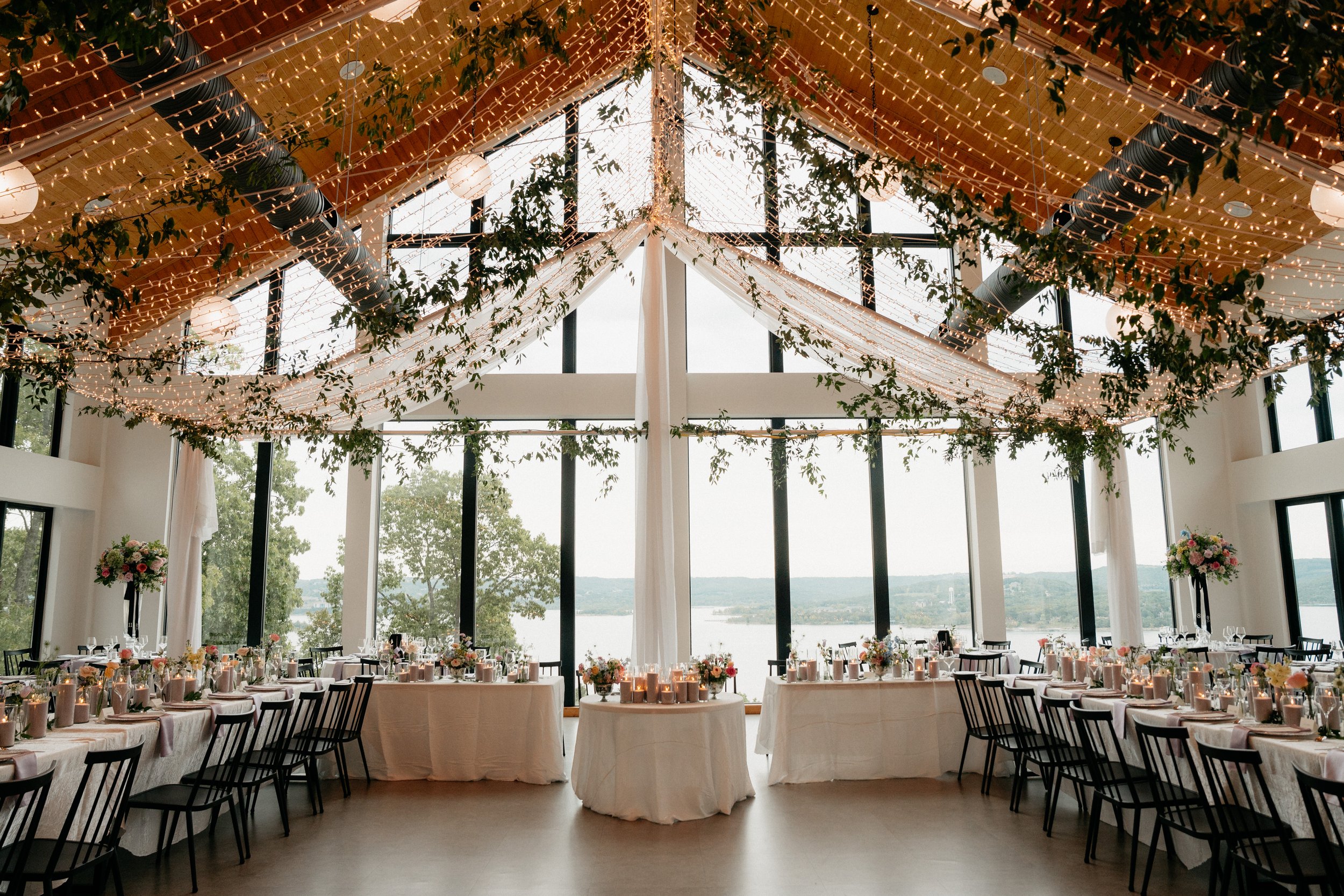 Elegant lakefront banquet hall decorated with string lights, hanging lanterns, and greenery. Large windows overlook Table Rock Lake, with tables set with flowers and candles for a celebration or wedding reception.