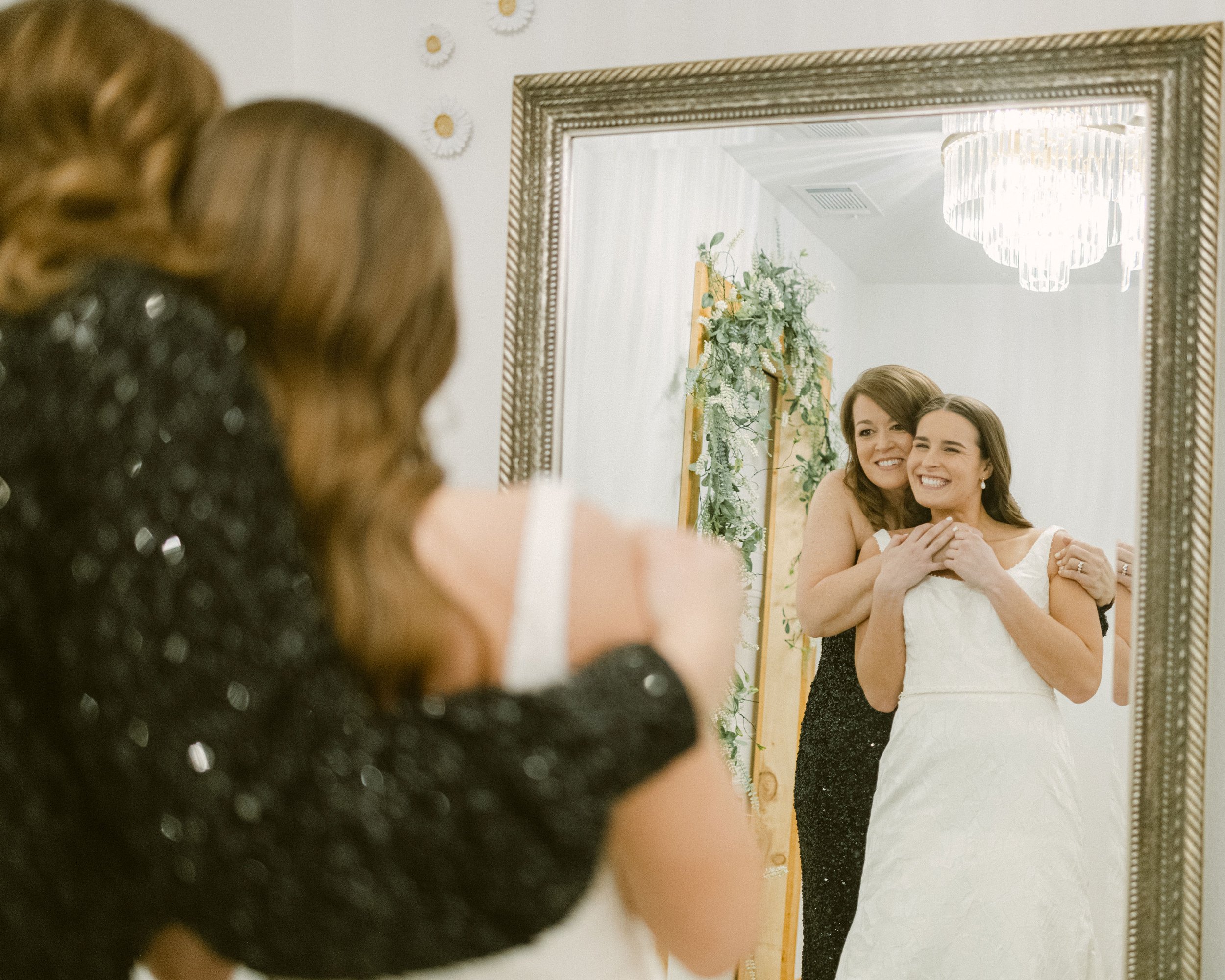 Two women, one in a wedding dress, smile and hug while looking at their reflection in a mirror.