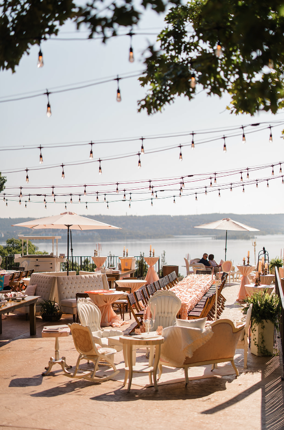 Outdoor wedding event with vintage furniture, string lights, umbrellas, and a view of a lake and trees in the background.