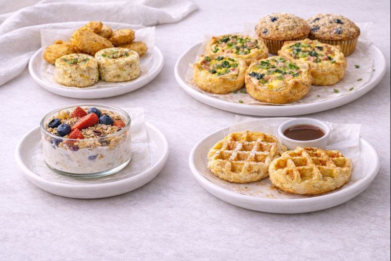 An assortment of breakfast items on white plates: hash brown patties with syrup, blueberry muffins and mini quiches, yogurt with berries and granola, and chicken nuggets with dipping sauce.