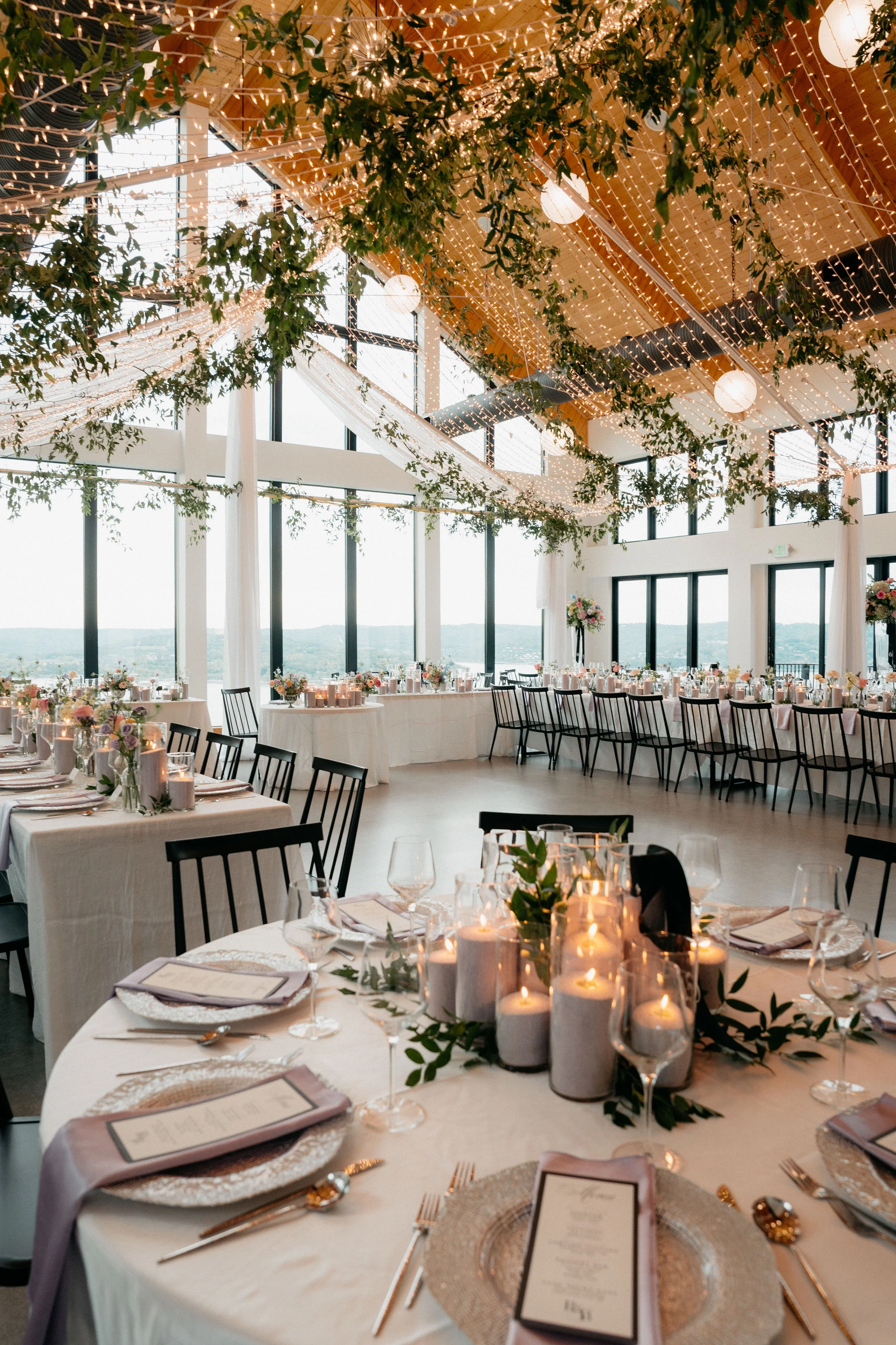 Elegant indoor wedding reception with round tables, floral centerpieces, candles, and string lights, featuring large windows and a scenic lake view outside.